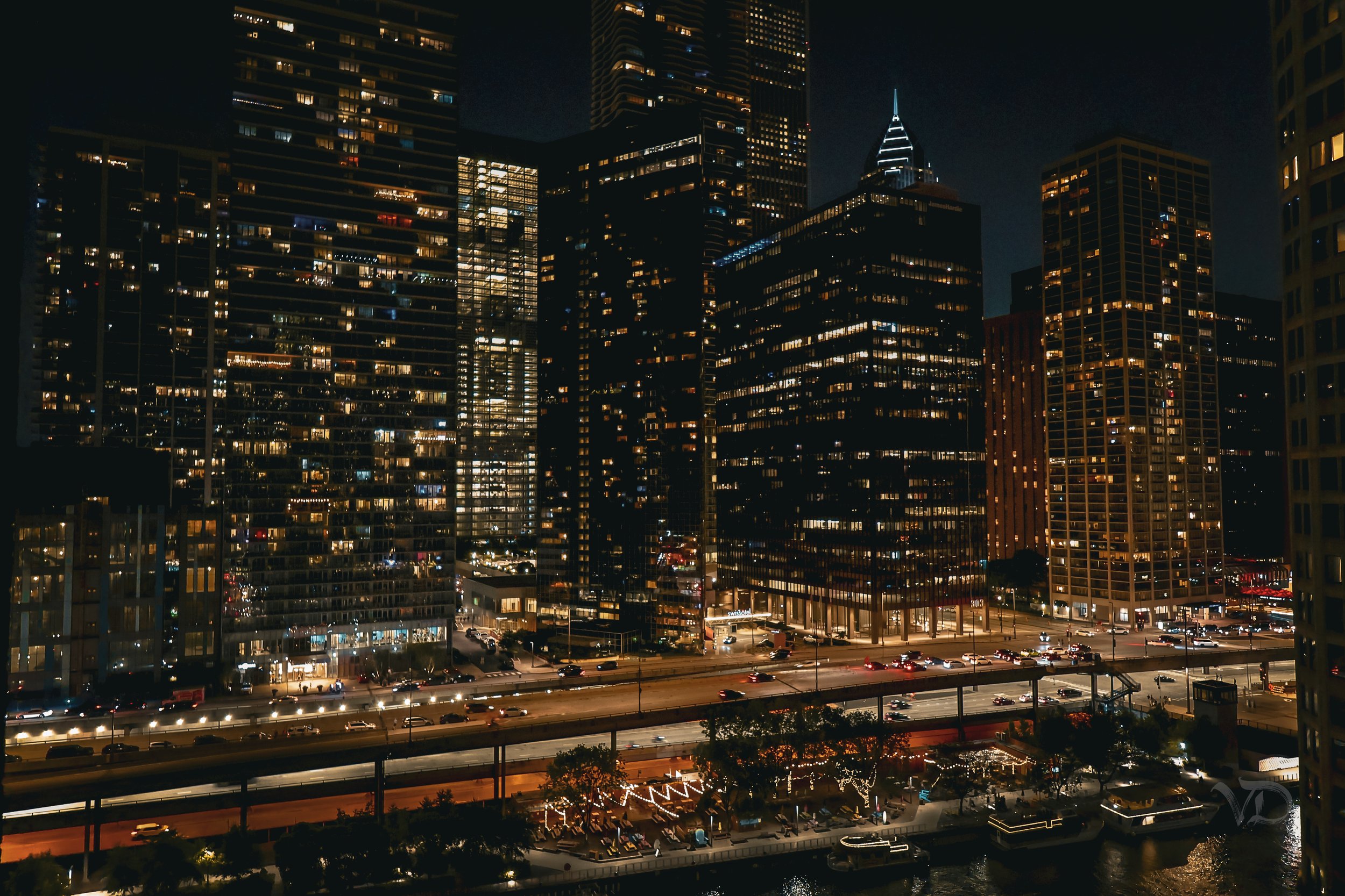 Nighttime cityscape with illuminated skyscrapers and a busy street with cars, a bridge, and lit outdoor seating area near the water.