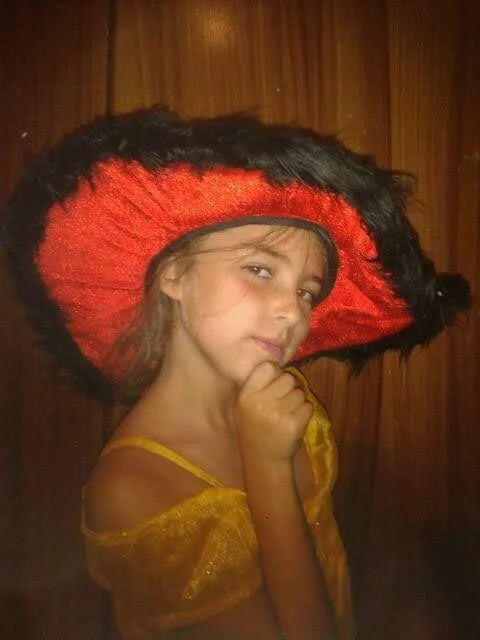 A young girl wearing a large red and black sombrero hat and a yellow top, standing in front of a wooden background, with her hand touching her chin and looking at the camera.
