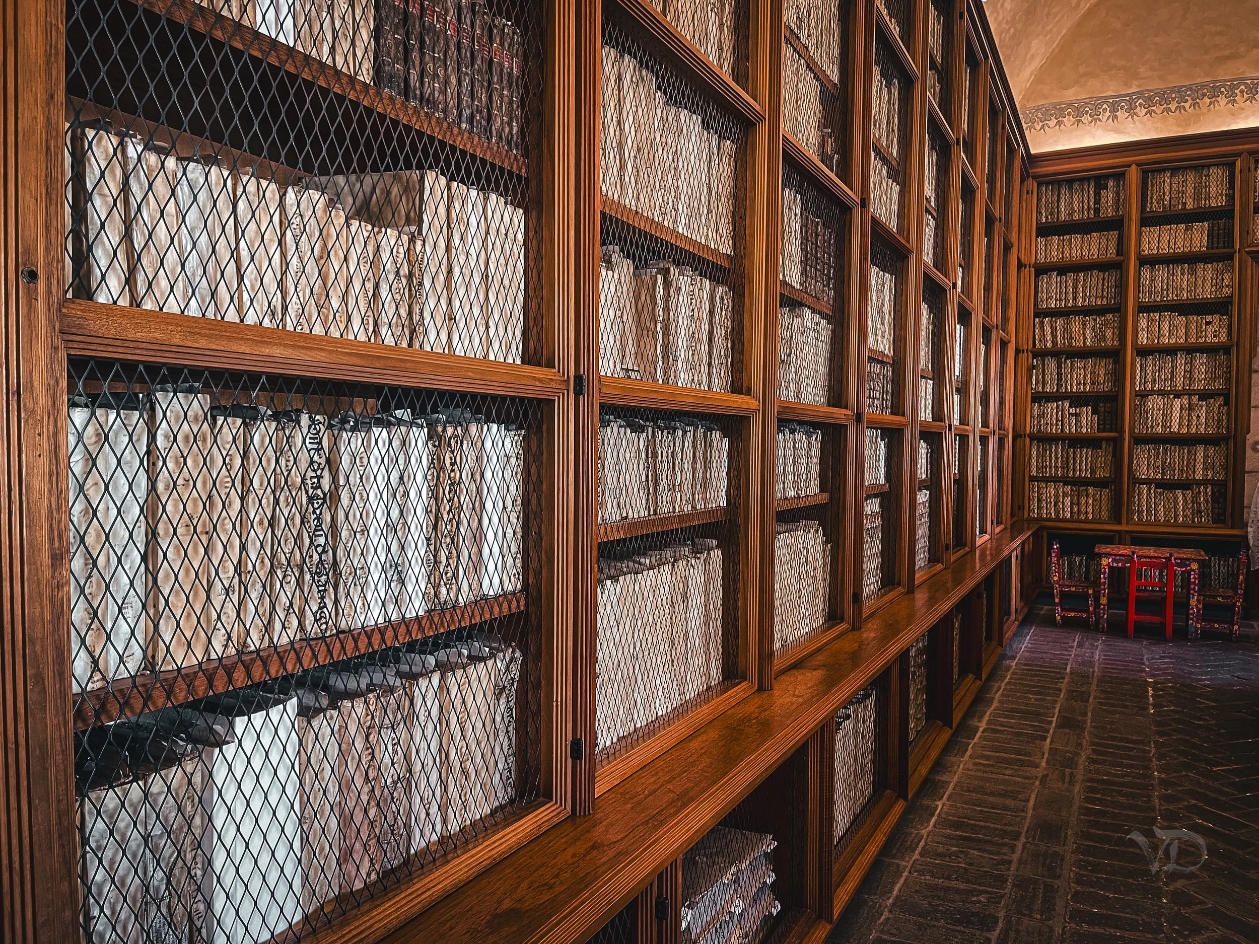 A large wooden library with a glass-fronted bookshelf filled with books, protected by a wire mesh. The room has dark wooden flooring and a small red table with chairs in the corner.