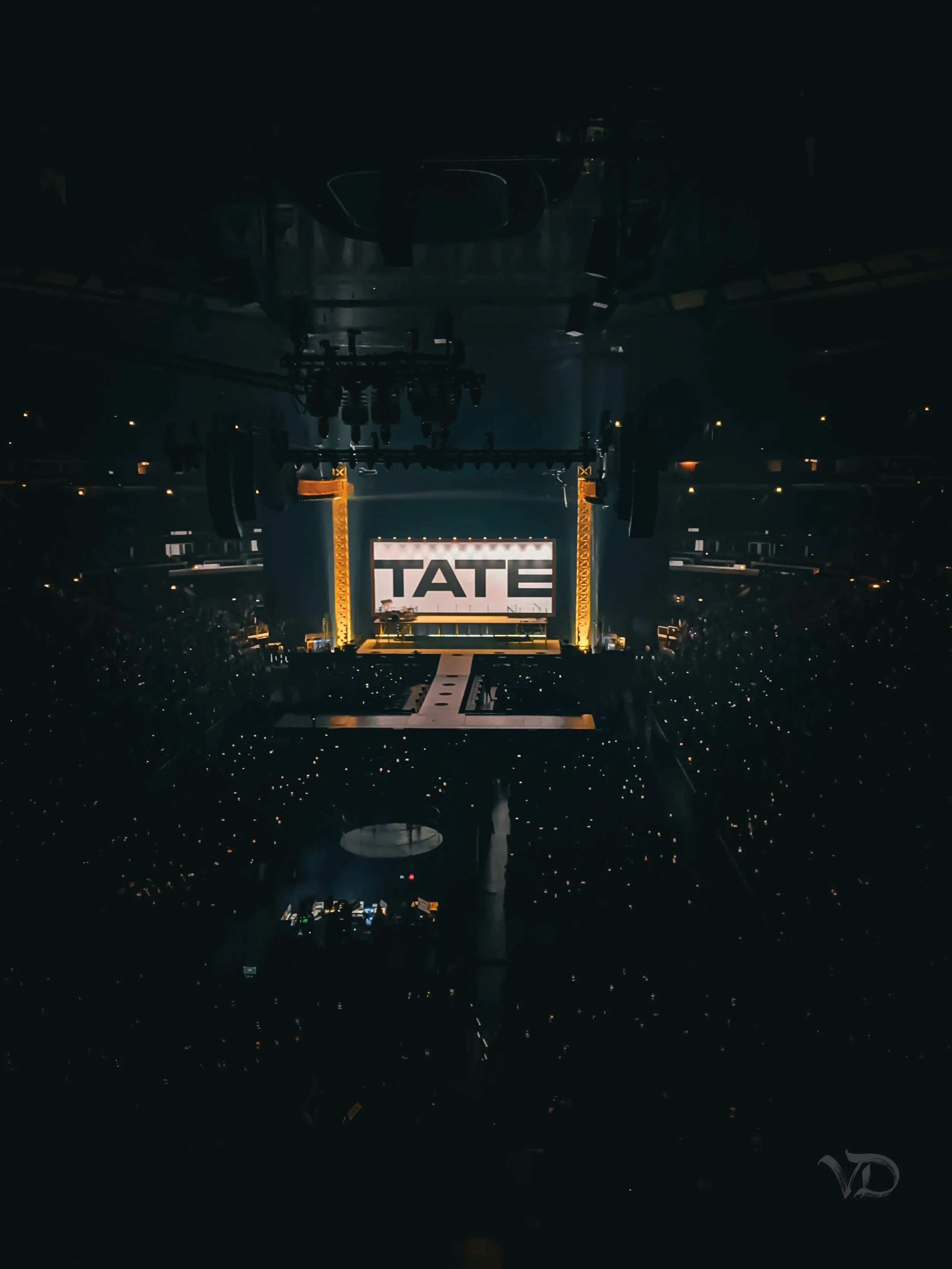 Stage with large screen displaying the word 'TATE' at a concert or event, surrounded by bright lights and an audience in a darkened venue.