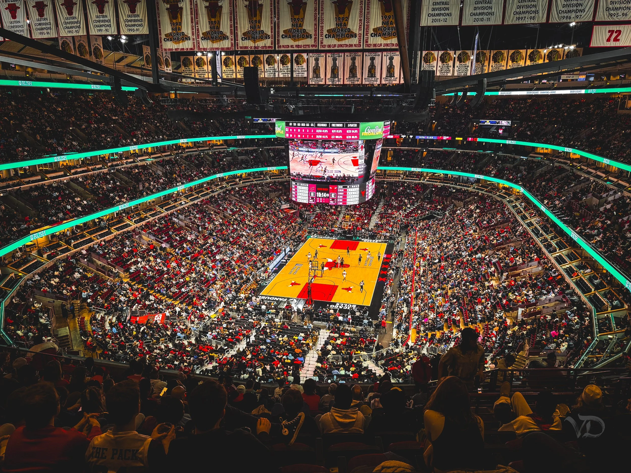 An indoor basketball arena filled with spectators, with a game in progress on the court. The court has a red logo at center, surrounded by advertising and a large video scoreboard hanging above.