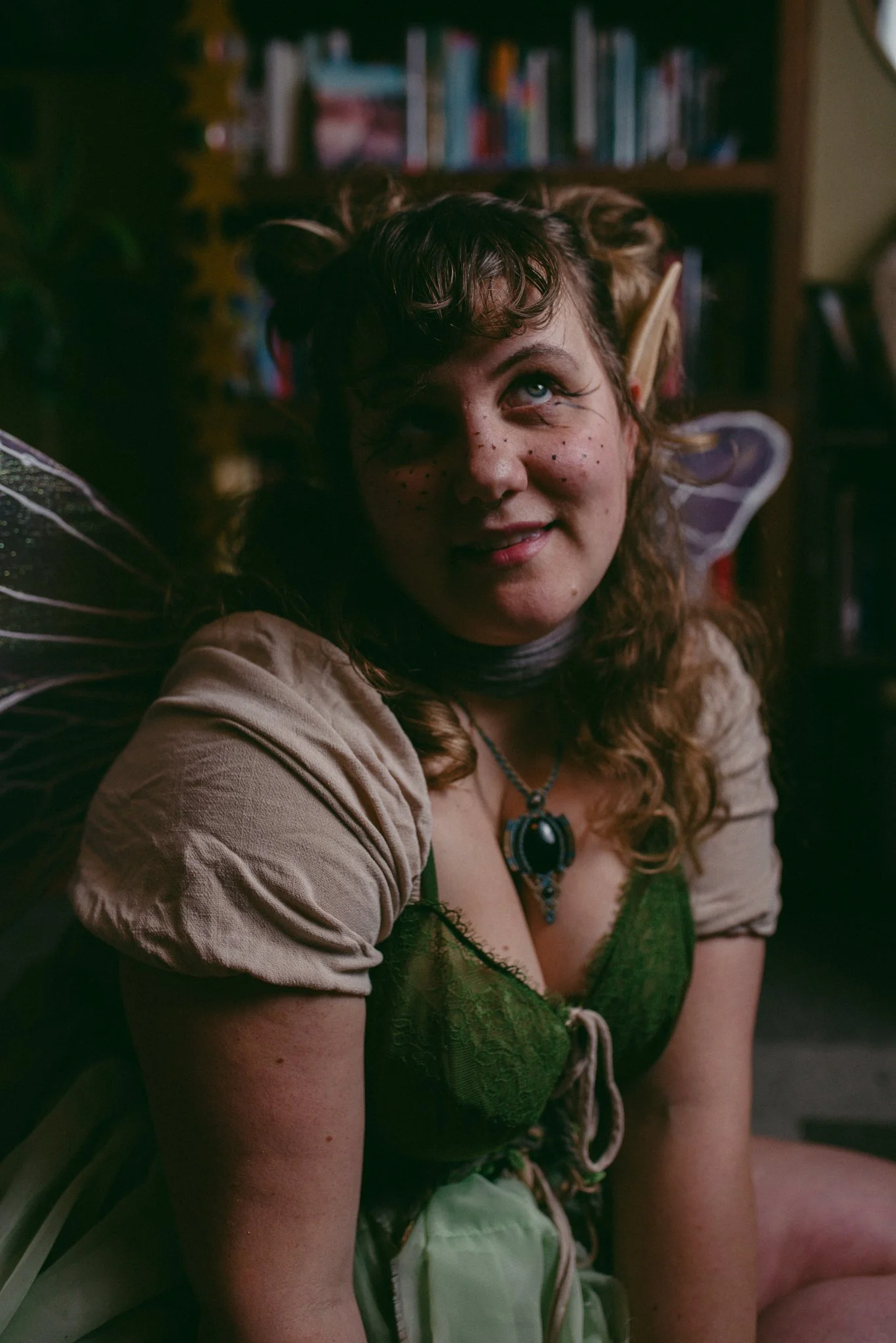 A woman dressed as a fairy with butterfly wings, curled hair, face freckles, and wearing a green top and necklace, smiling indoors with books in the background.