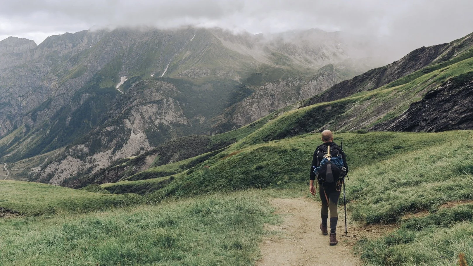 A hiker with a backpack and walking stick hiking along a dirt trail through green grassy mountains with cloudy skies.