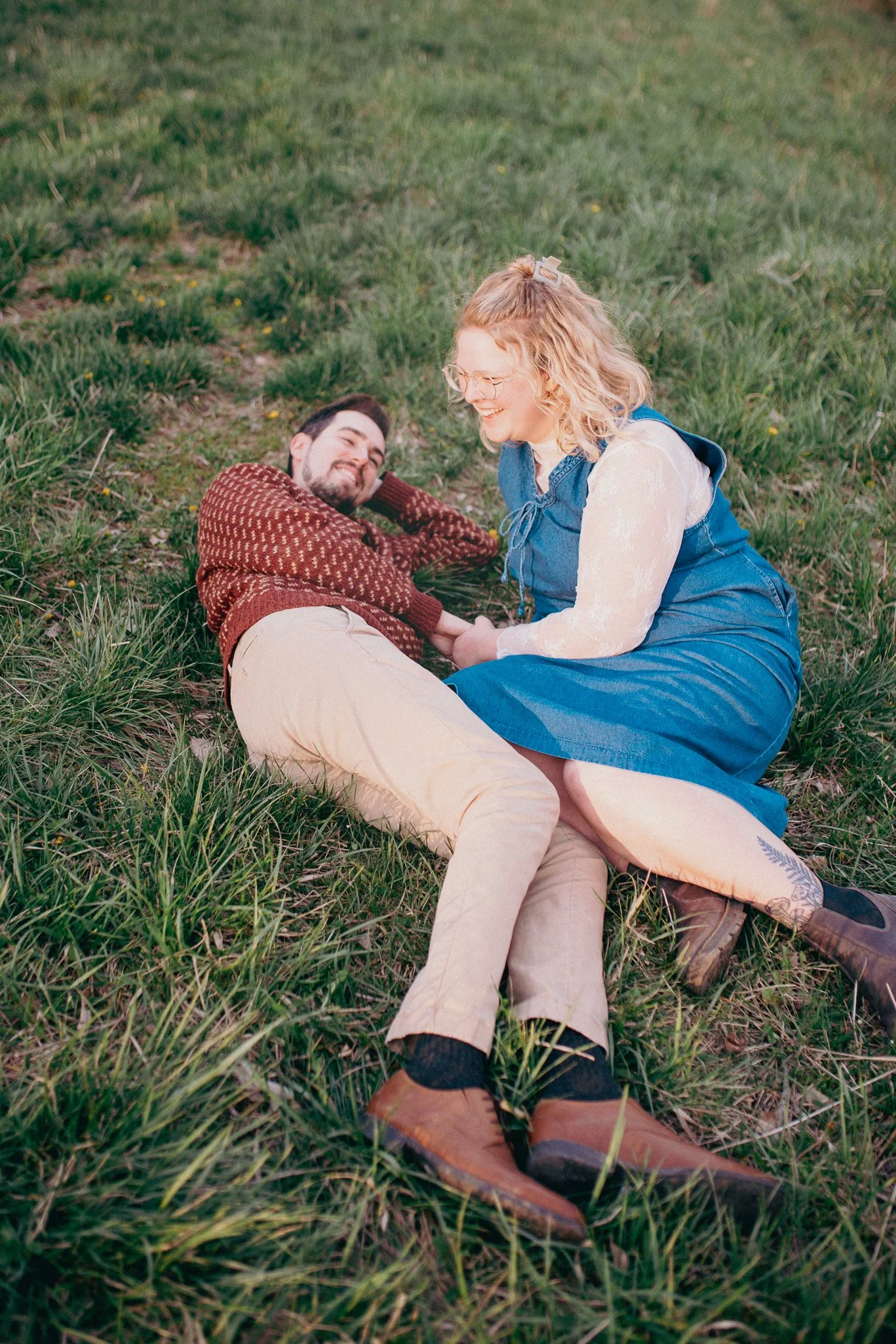 Engagement Photoshoot of a man and a woman lying in the grass, smiling and holding hands, with the woman sitting and the man reclining. Richmond, Indiana.