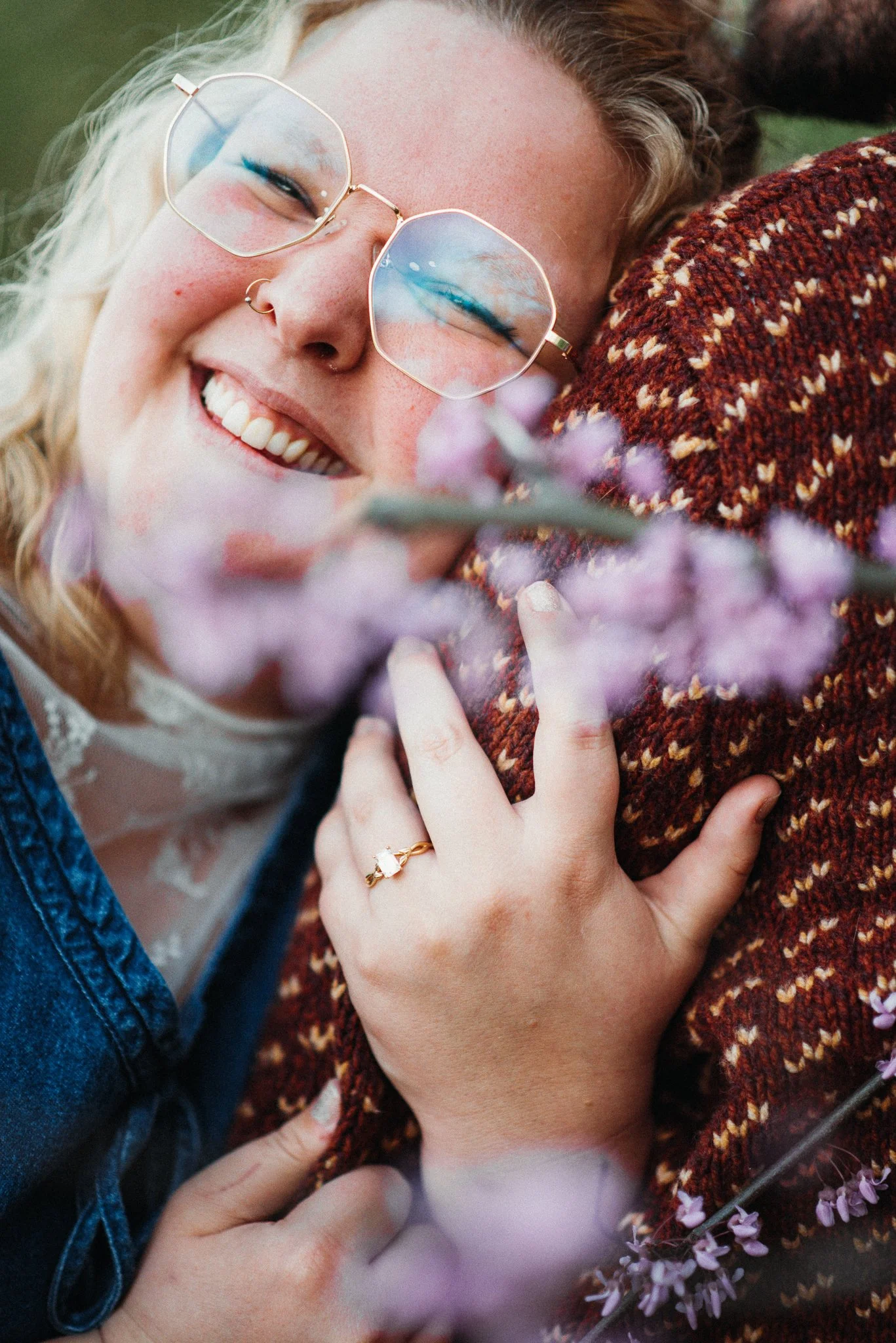 Engagement photoshoot of woman holder her fiancé's arm with engagement ring on, standing in front of red bud blossoms. Richmond, Indiana.