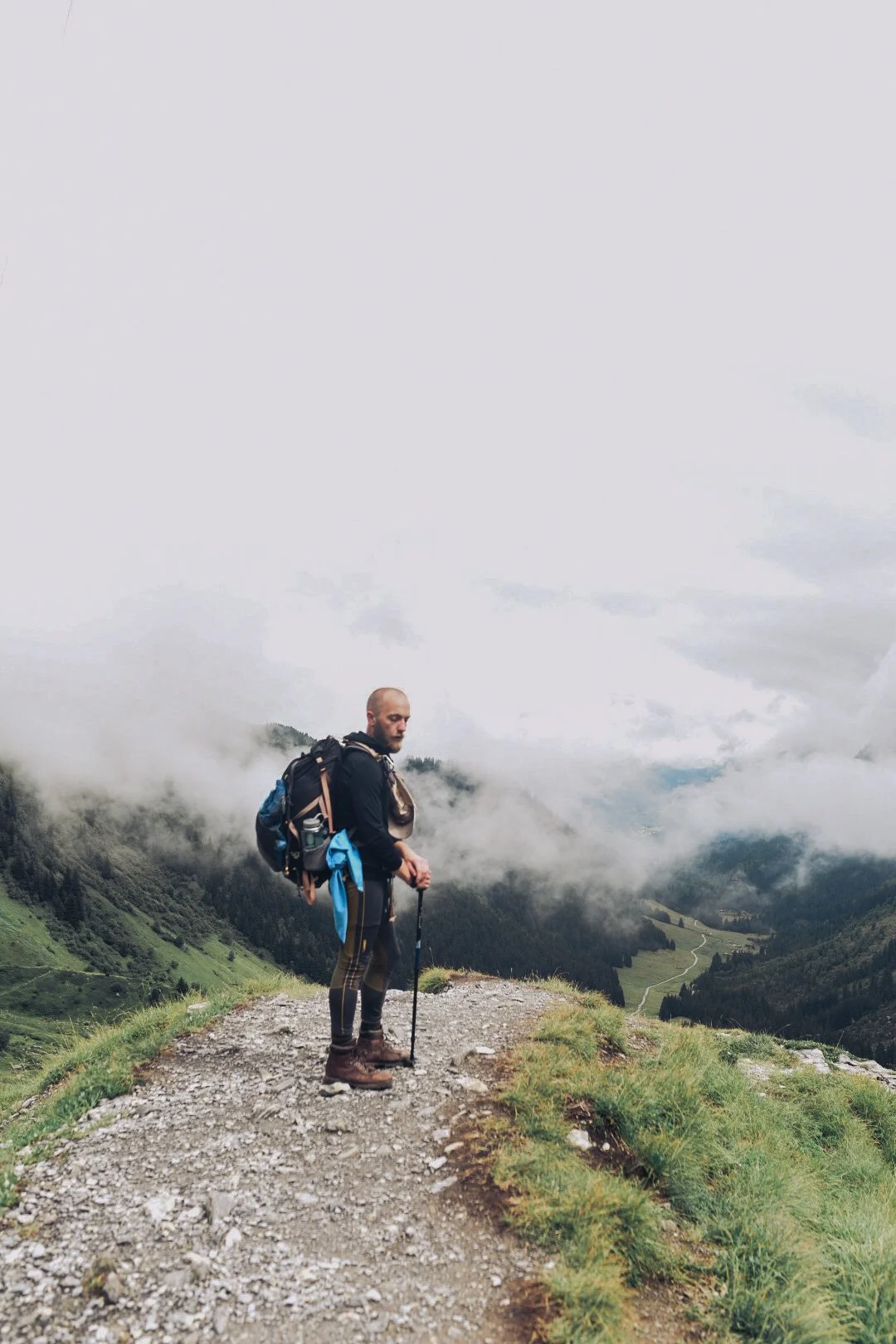 A man standing with a hiking pole on a mountain trail overlooking a foggy valley and green hills.