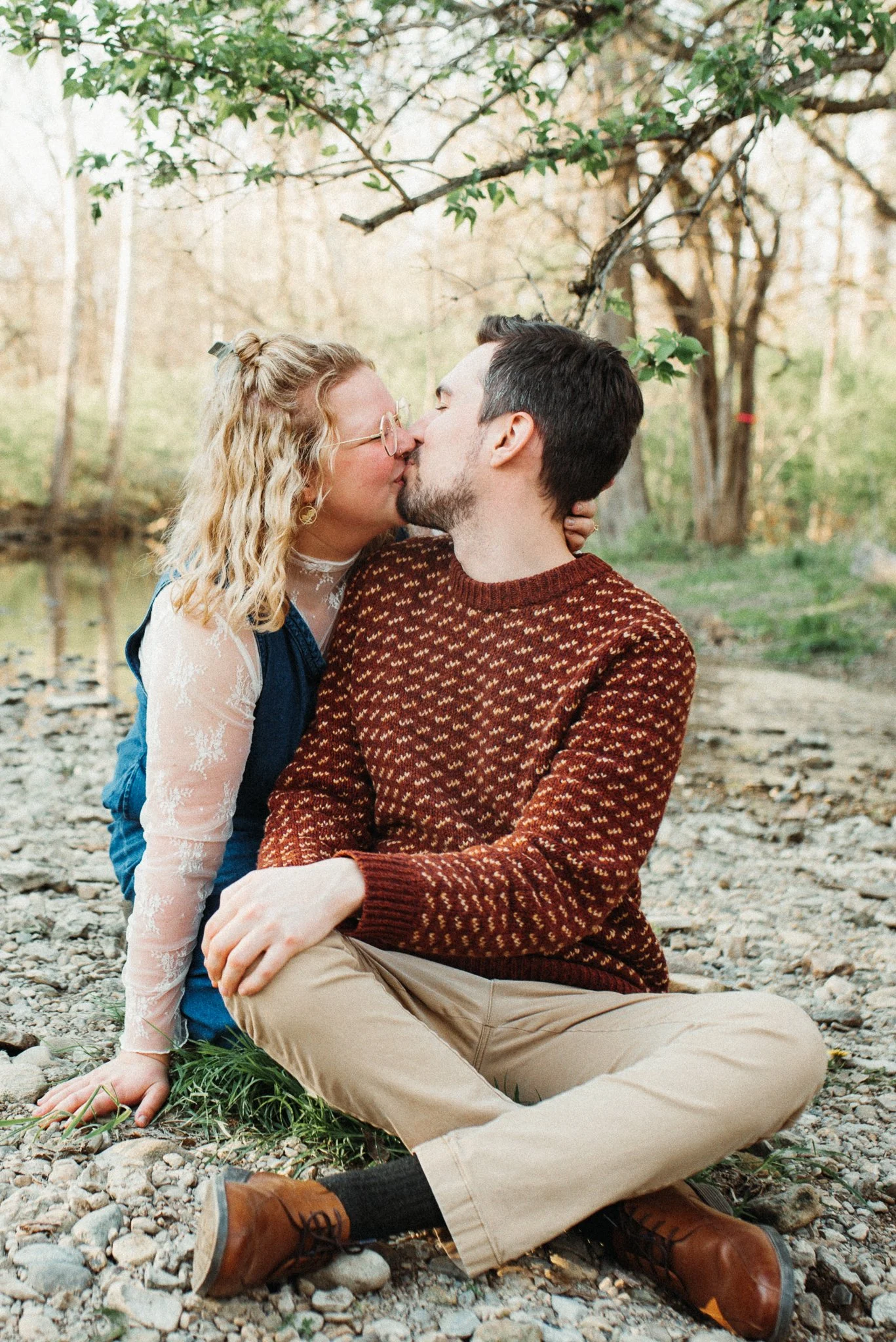 Engagement Photoshoot of a couple kissing outdoors during daytime, sitting near a creek surrounded by trees with Spring leaves. Richmond, Indiana.
