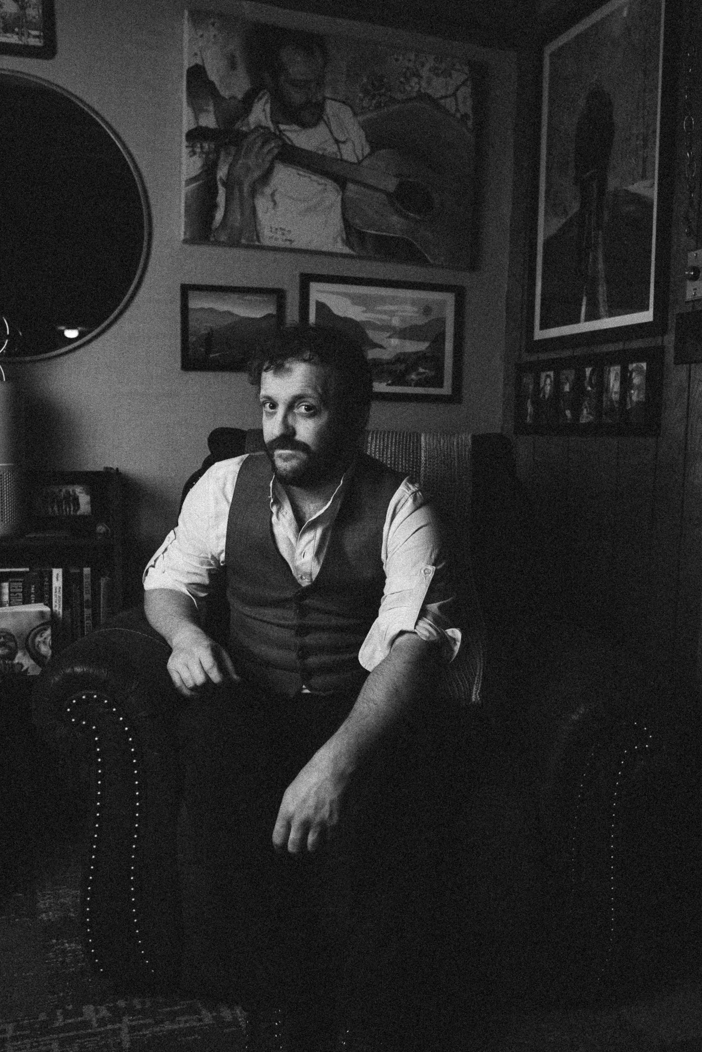 A man with a beard and mustache sitting in a vintage armchair in a room filled with framed artwork, books, and a guitar. The photo is in black and white.