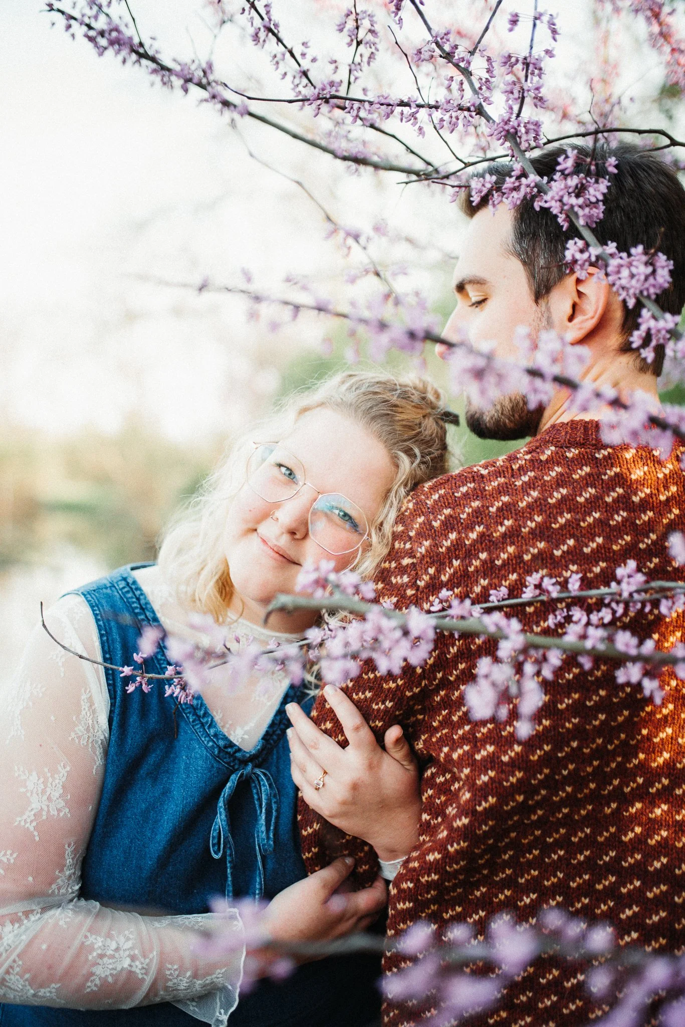 Engagement photoshoot of woman holder her fiancé's arm with engagement ring on, standing in front of red bud blossoms. Richmond, Indiana.