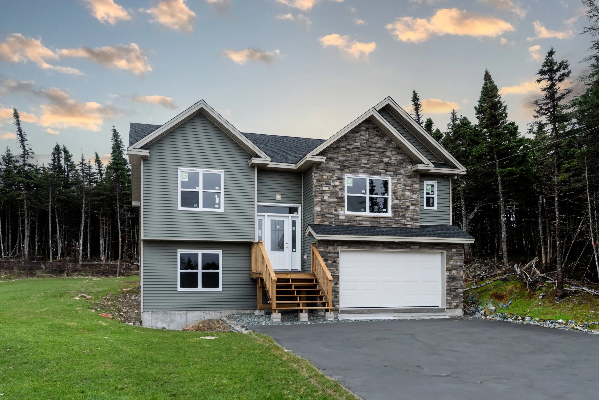 Newly constructed two-story house with gray siding, stone accents, white garage door, and a small stairway leading to the front door, set against a backdrop of pine trees and a partly cloudy sky at sunset. New Home Built by Viking Construction in New