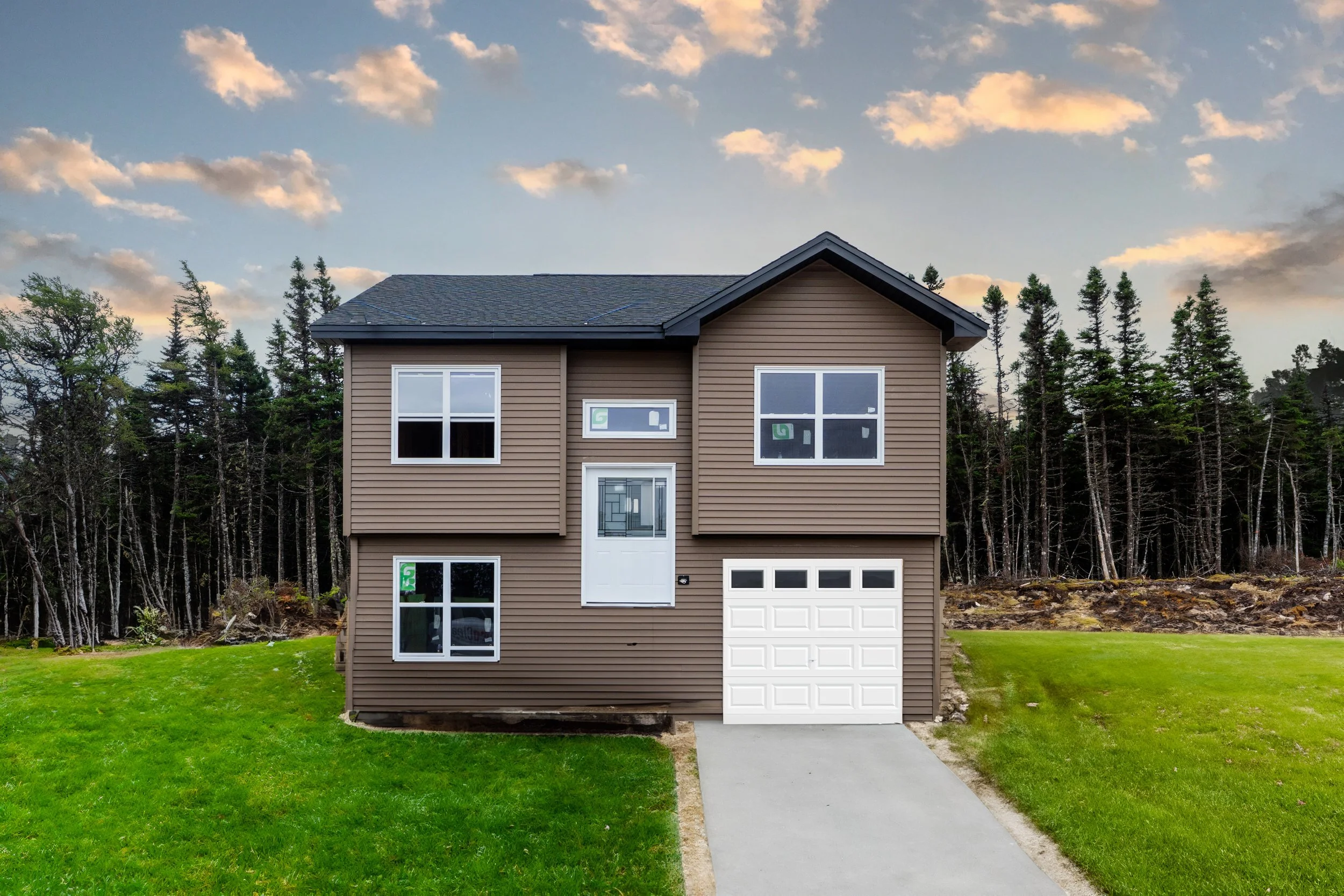 Newly constructed two-story house with brown siding, white garage door, and multiple large windows, situated on a grassy lot with a forested area in the background, under a partly cloudy sky. New Home Built by Viking Construction in Newfoundland.