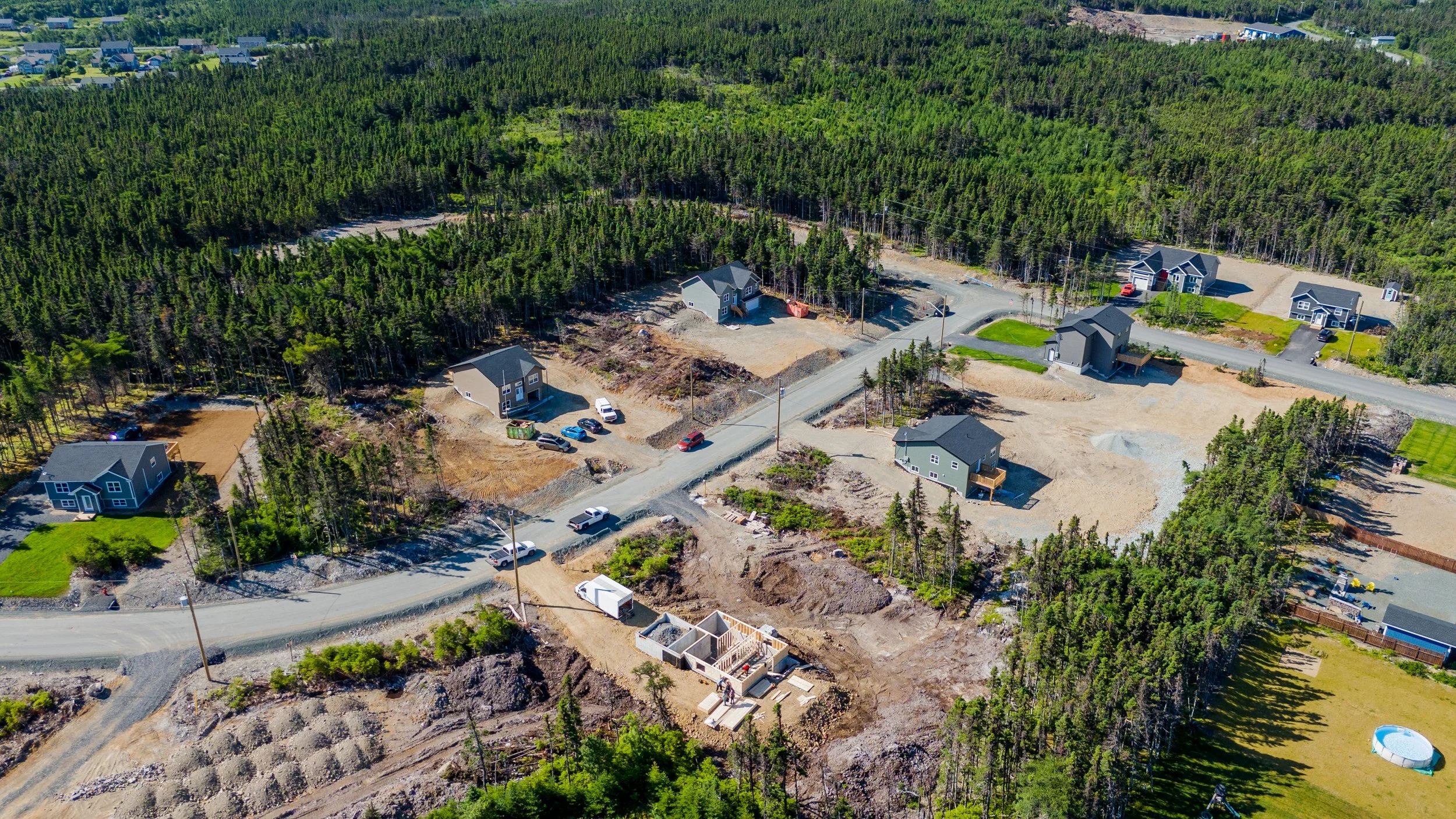 Aerial view of a residential construction site with houses in various stages of building, surrounded by a dense forest and some completed homes. in Newfoundland's newest subdivision of Spruce Grove