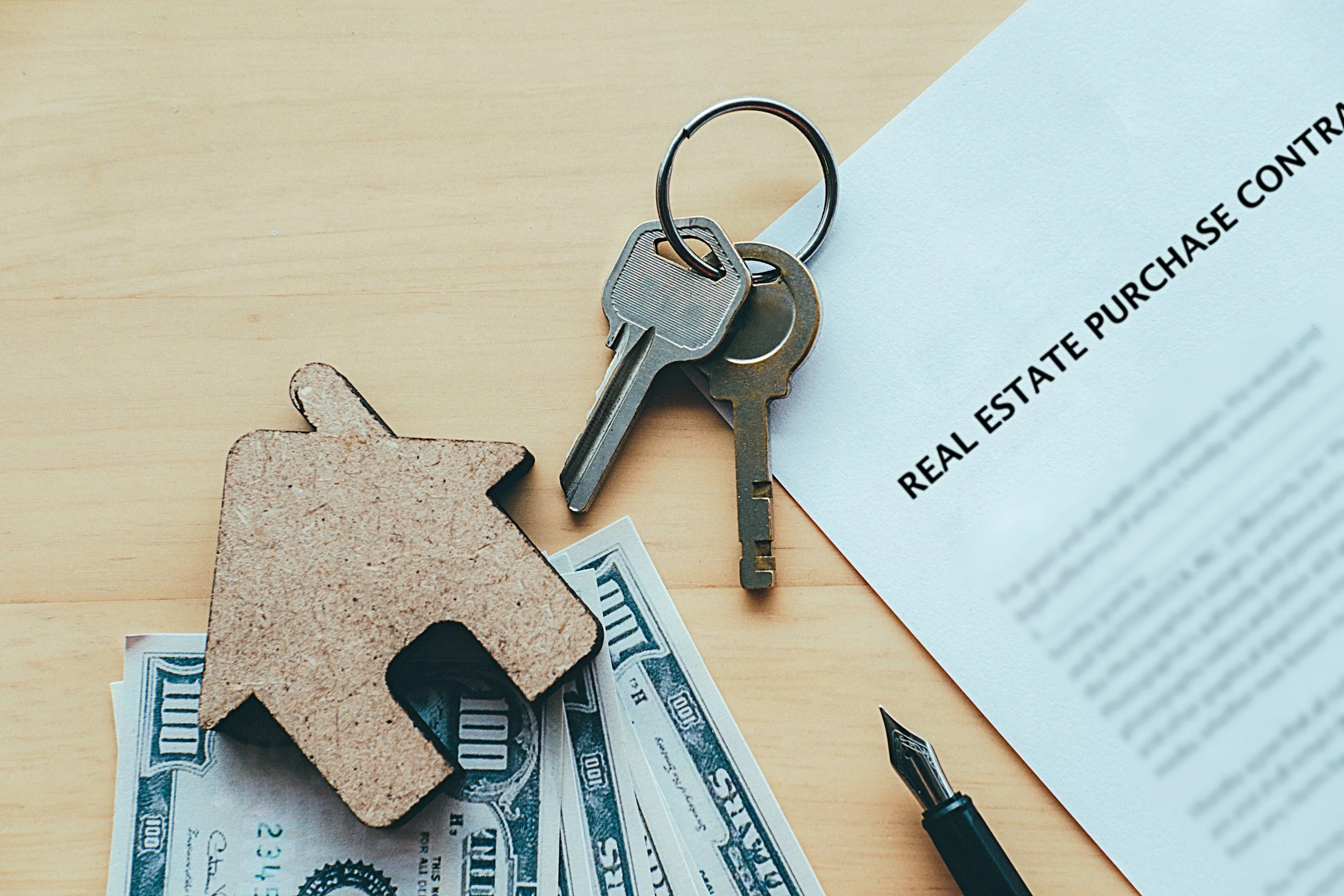 A set of keys, a small house-shaped cutout, U.S. dollar bills, a real estate purchase contract, and a fountain pen on a wooden surface.