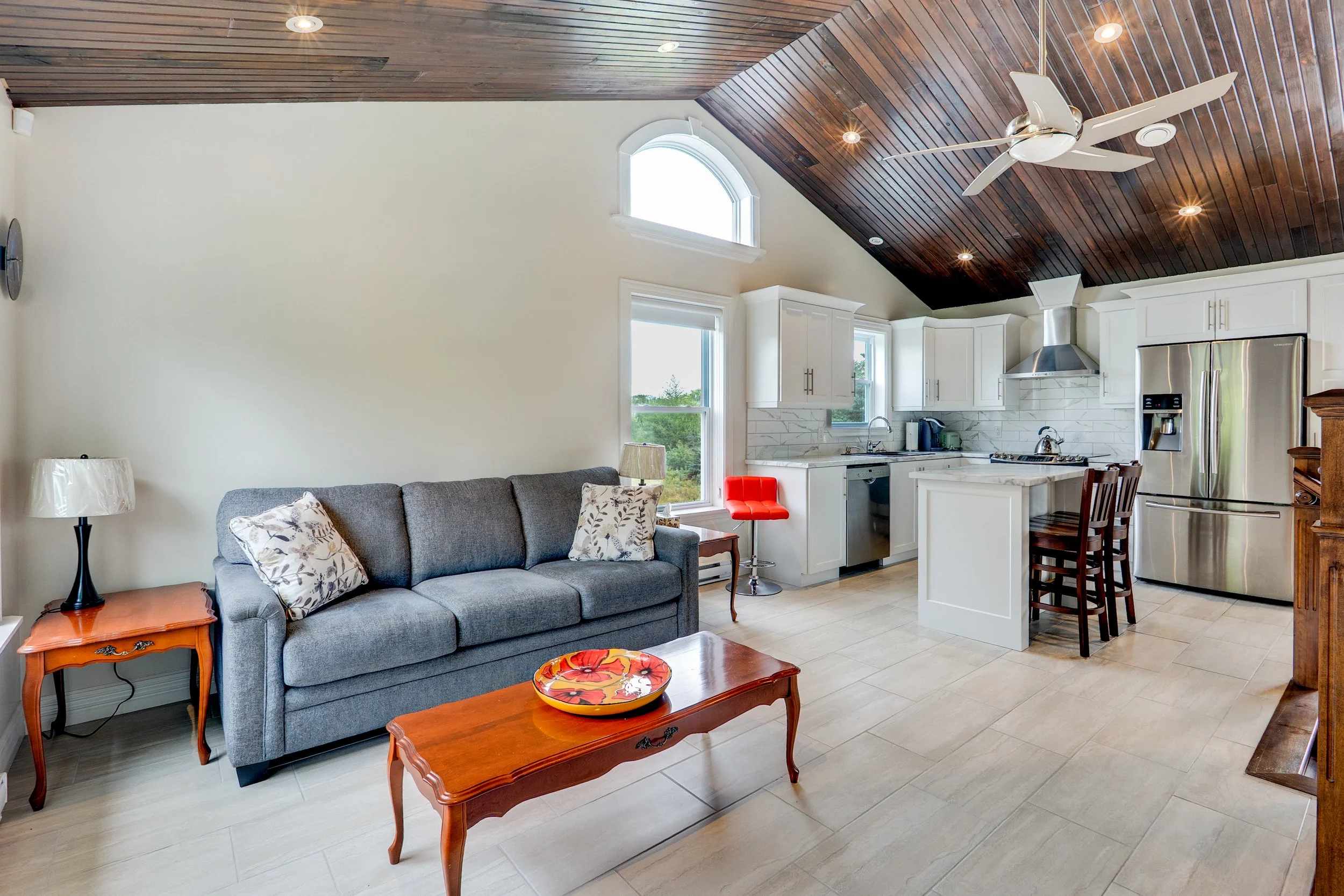 Open-concept living room and kitchen with white cabinets, stainless steel refrigerator, and dark wood ceiling with ceiling fan.