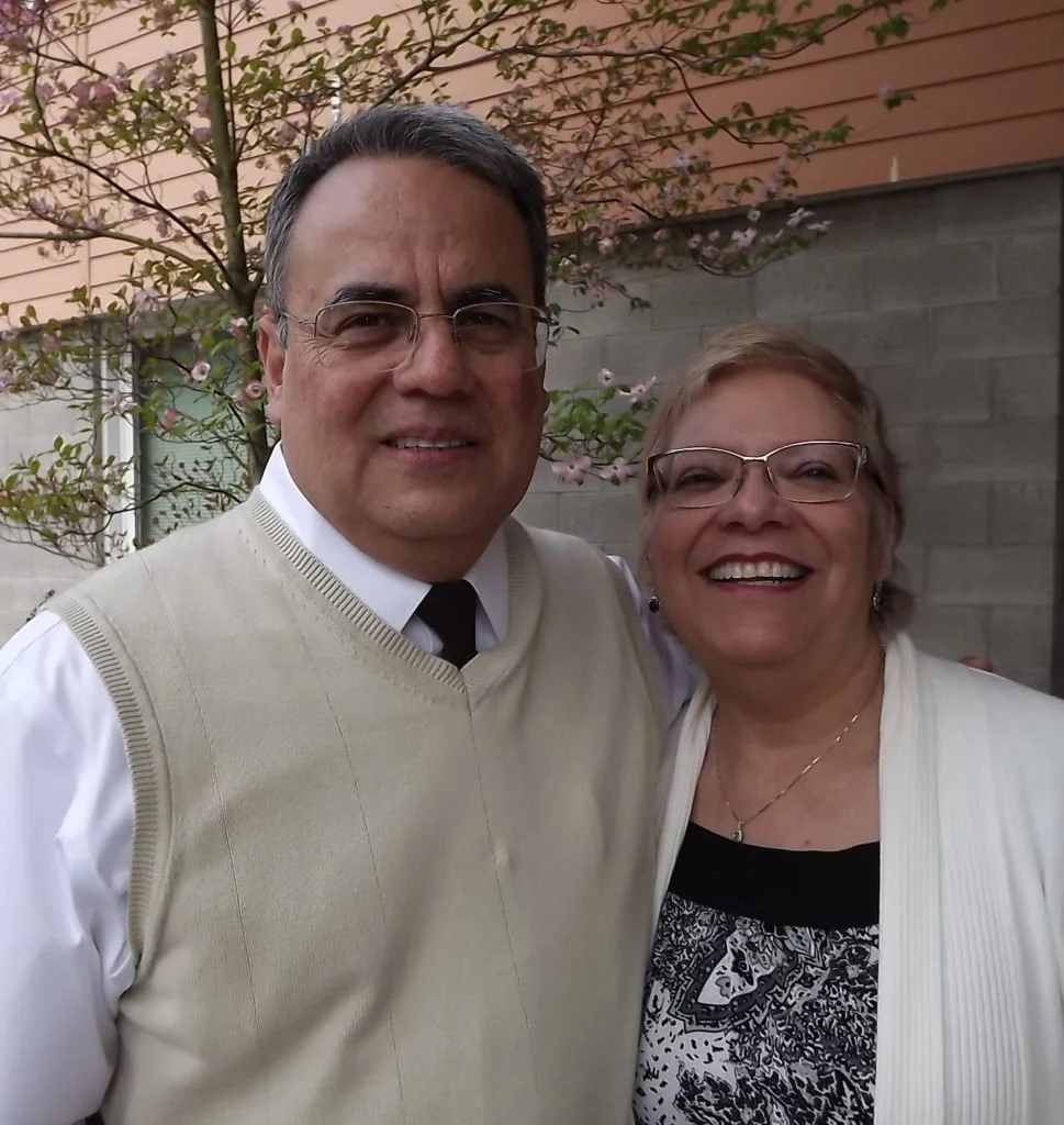A man and woman smiling outdoors, standing close together in front of a tree with pink flowers and a brick wall.