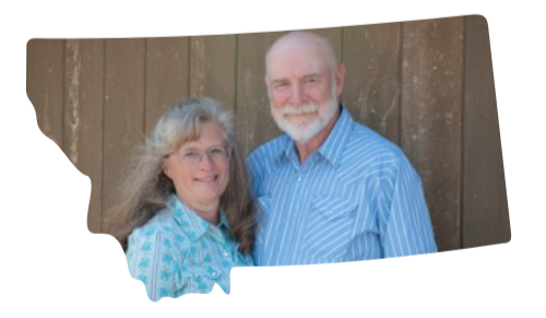 A smiling white-haired couple standing outdoors in front of a wooden fence, both wearing blue patterned shirts.