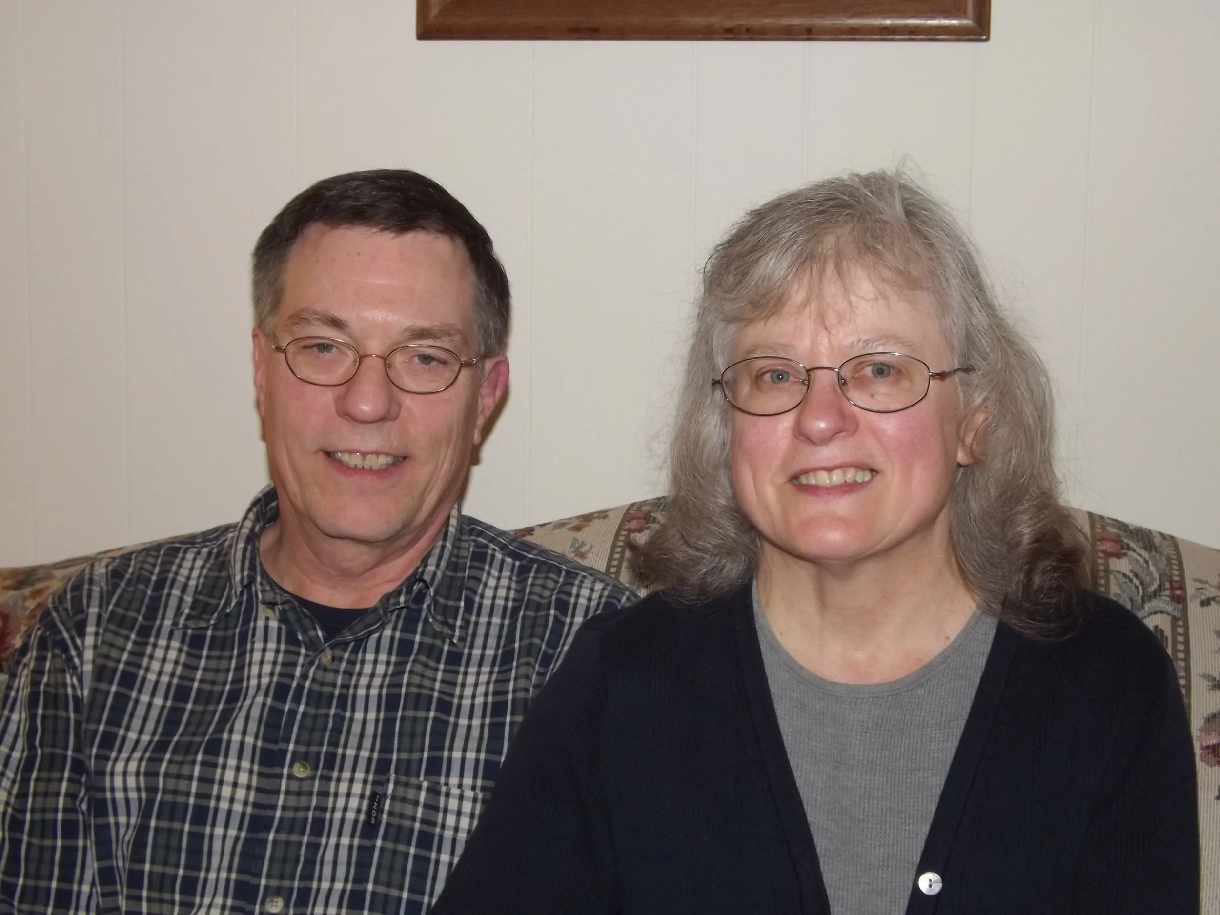 A man and woman sitting on a floral-patterned sofa in front of a beige wall with a wooden-framed picture hanging above them. The man has short dark hair, glasses, and is wearing a checkered shirt. The woman has shoulder-length gray hair, glasses, and is wearing a black cardigan over a gray top.