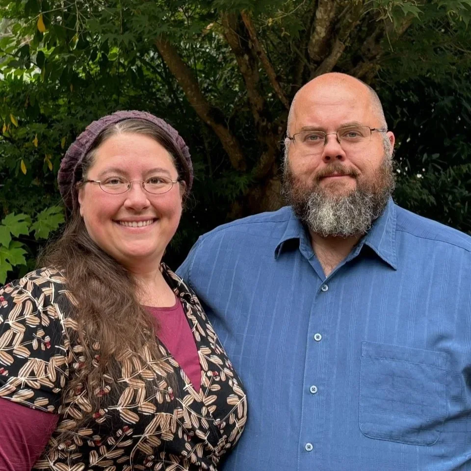 A smiling woman with glasses and long brown hair, wearing a patterned jacket and a maroon shirt, standing next to a serious-looking man with glasses, a bald head, and a full gray beard, dressed in a blue shirt, outdoors in front of green foliage.