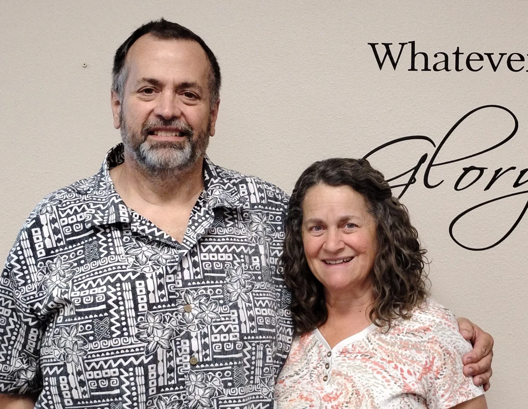 A man and woman standing close together, smiling in front of a wall with the word 'Glory' written on it.