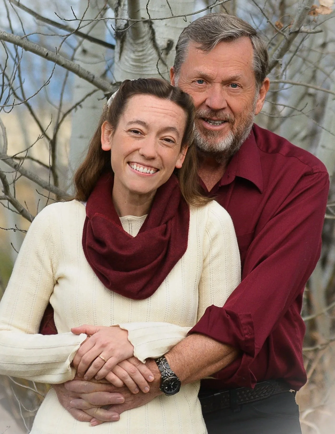 A smiling couple outdoors in front of bare trees, woman wearing a cream sweater and burgundy scarf, man in a burgundy shirt.