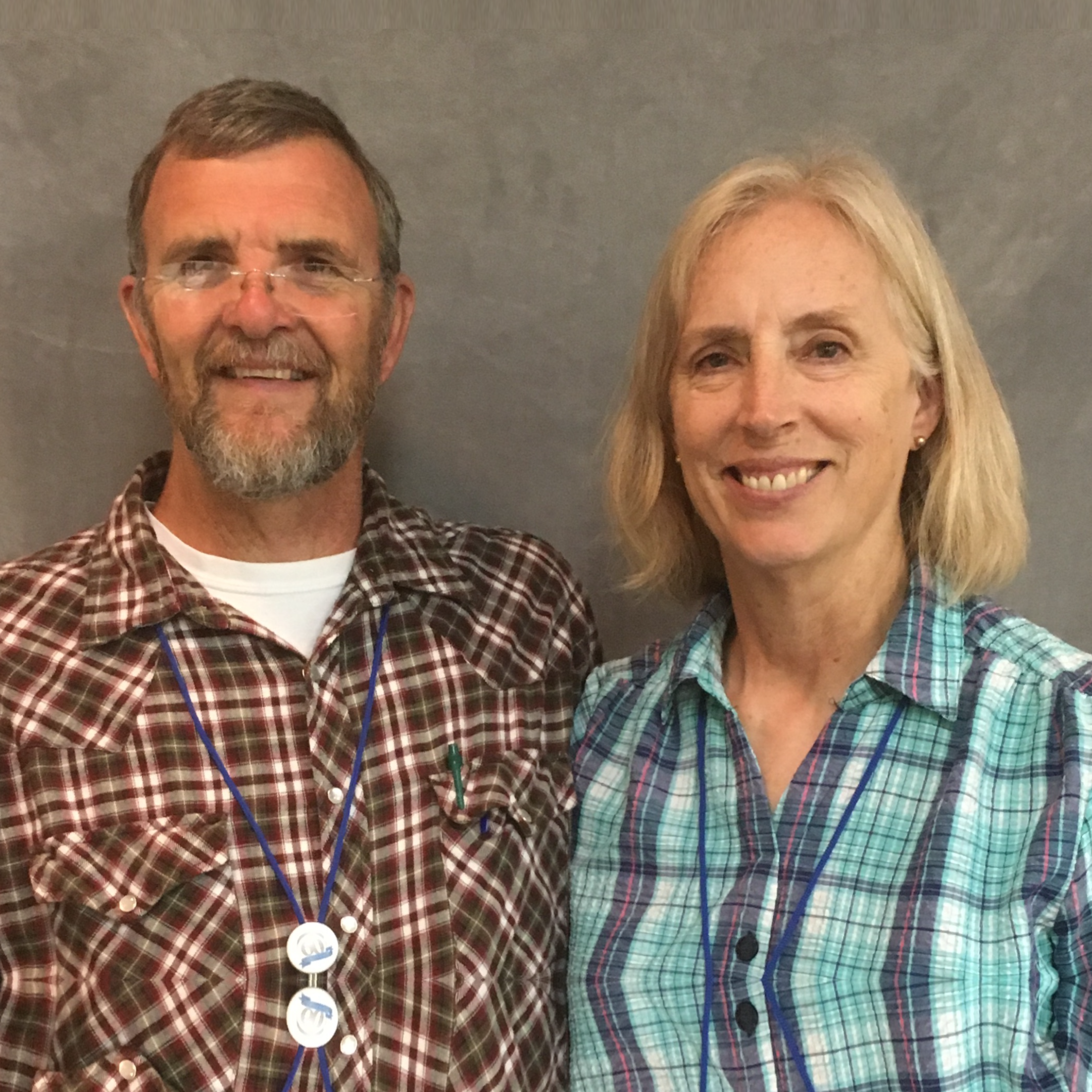 A smiling man with a beard and glasses standing next to a smiling woman with shoulder-length blonde hair, both wearing plaid shirts and blue lanyards, against a plain gray background.