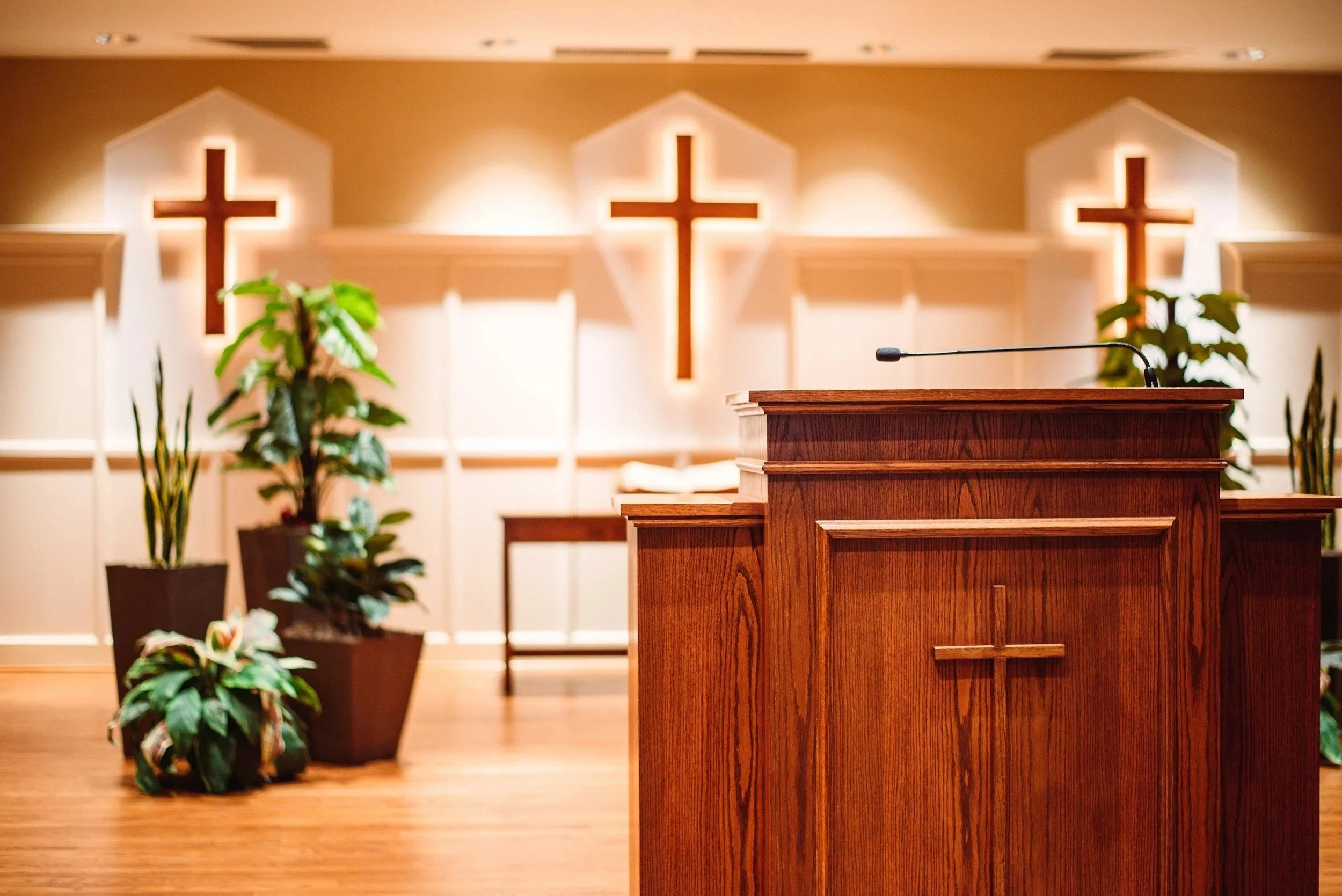 A church interior with a wooden pulpit featuring a cross, surrounded by potted plants, and illuminated crosses on the wall in the background.
