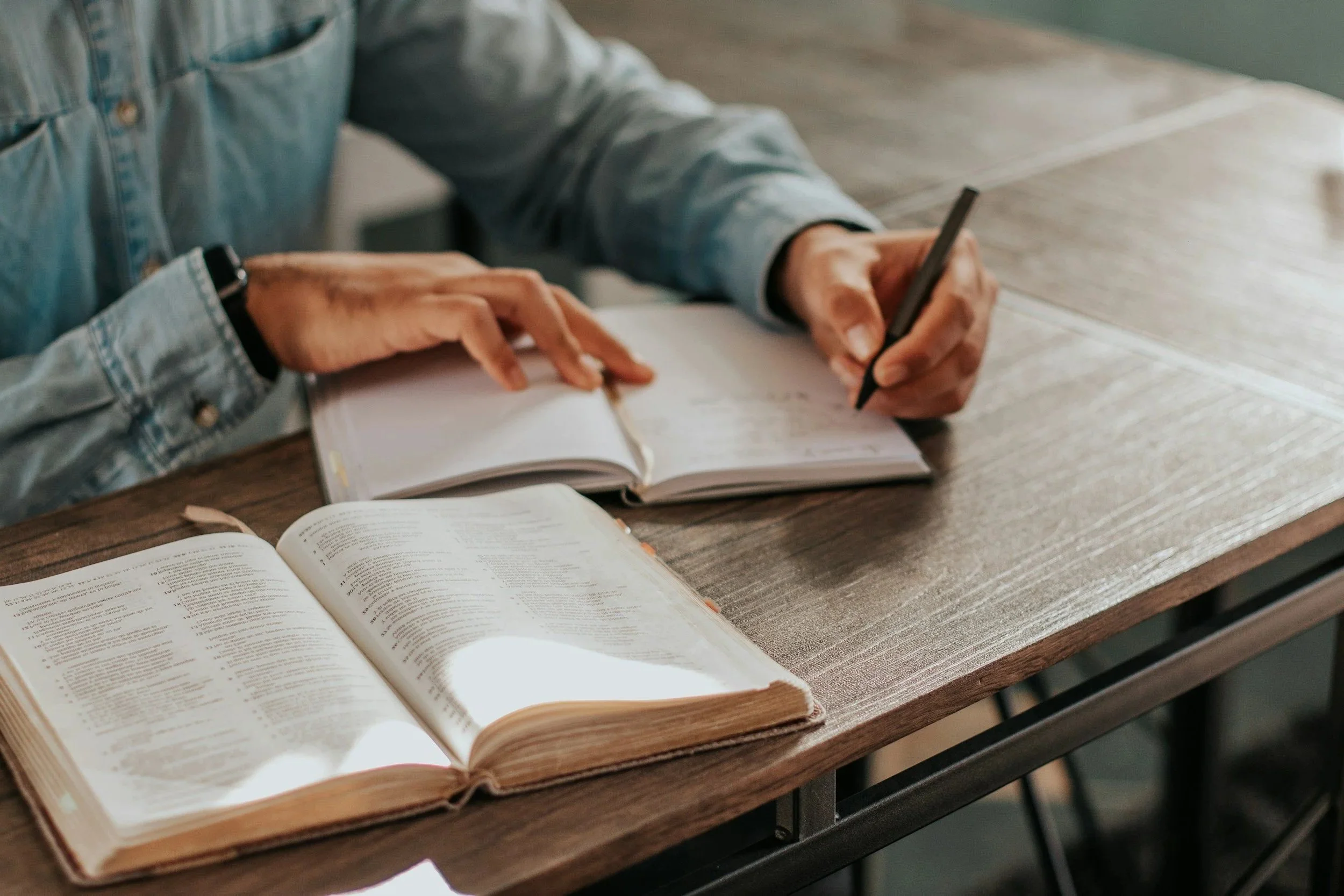 Person writing in a notebook at a wooden table, with an open book in front of them.