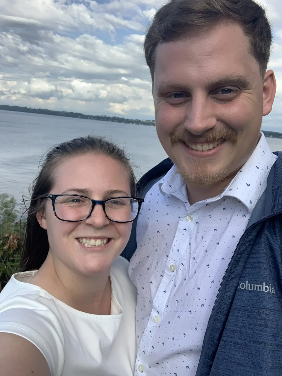 A smiling couple taking a selfie beside a body of water under a partly cloudy sky.
