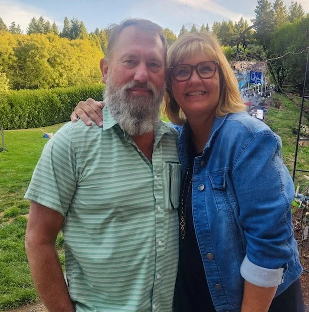 A man and woman smiling outdoors with trees and a green lawn in the background, standing close together with the woman’s arm around the man's shoulder.