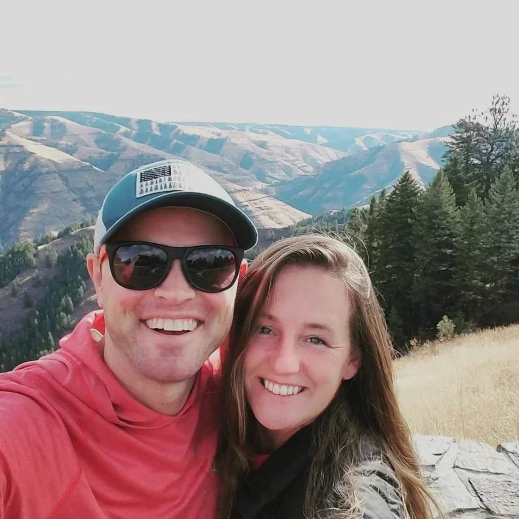 Smiling couple taking selfie outdoors with a mountain landscape and forest in the background.