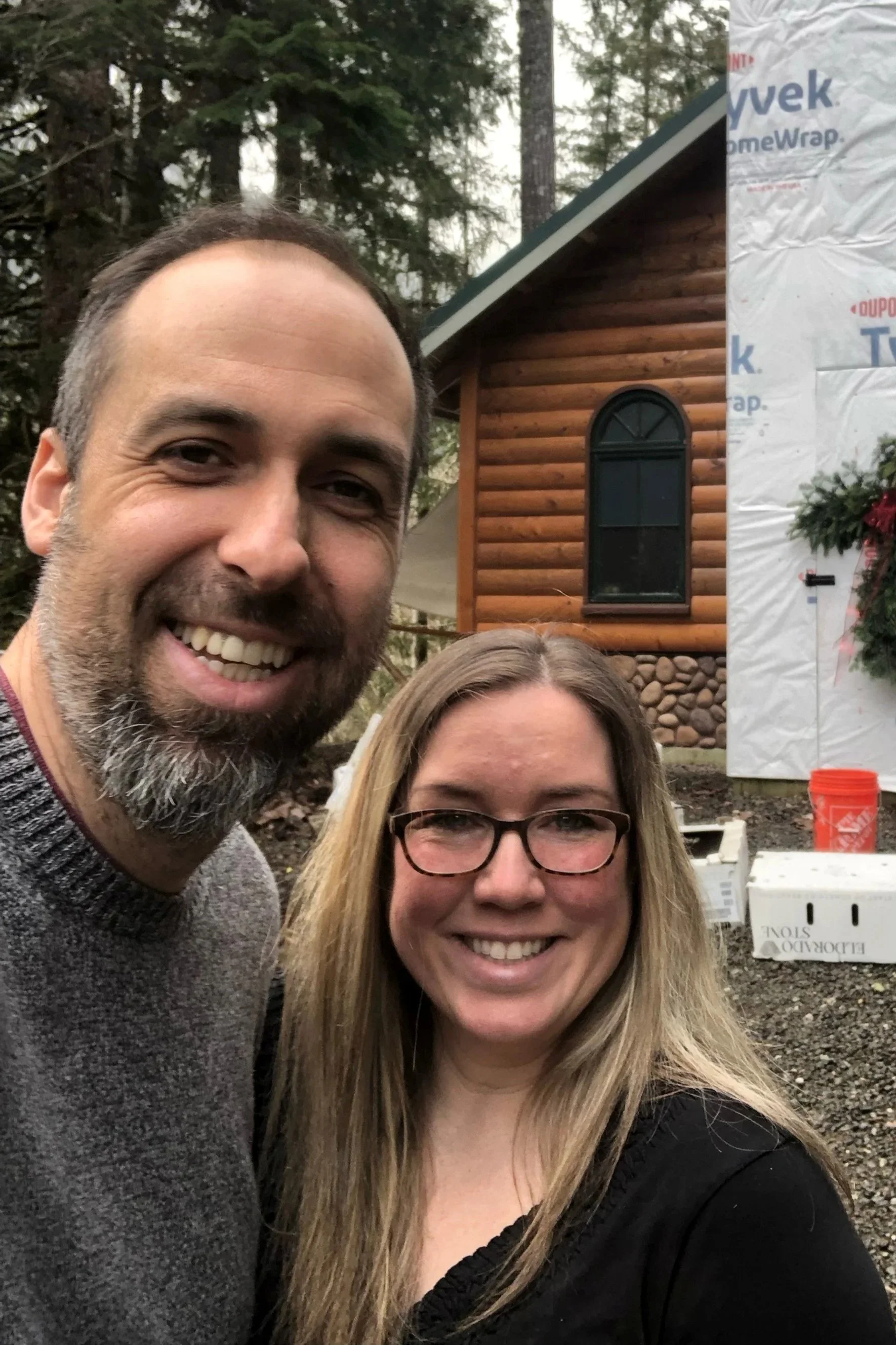 A smiling man and woman taking a selfie in front of a partially built wooden house with a brick foundation, surrounded by trees.