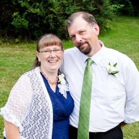 A smiling woman and a smiling man standing outdoors on grass with trees in the background, dressed in semi-formal attire with boutonniere and corsage, possibly at a wedding or special occasion.