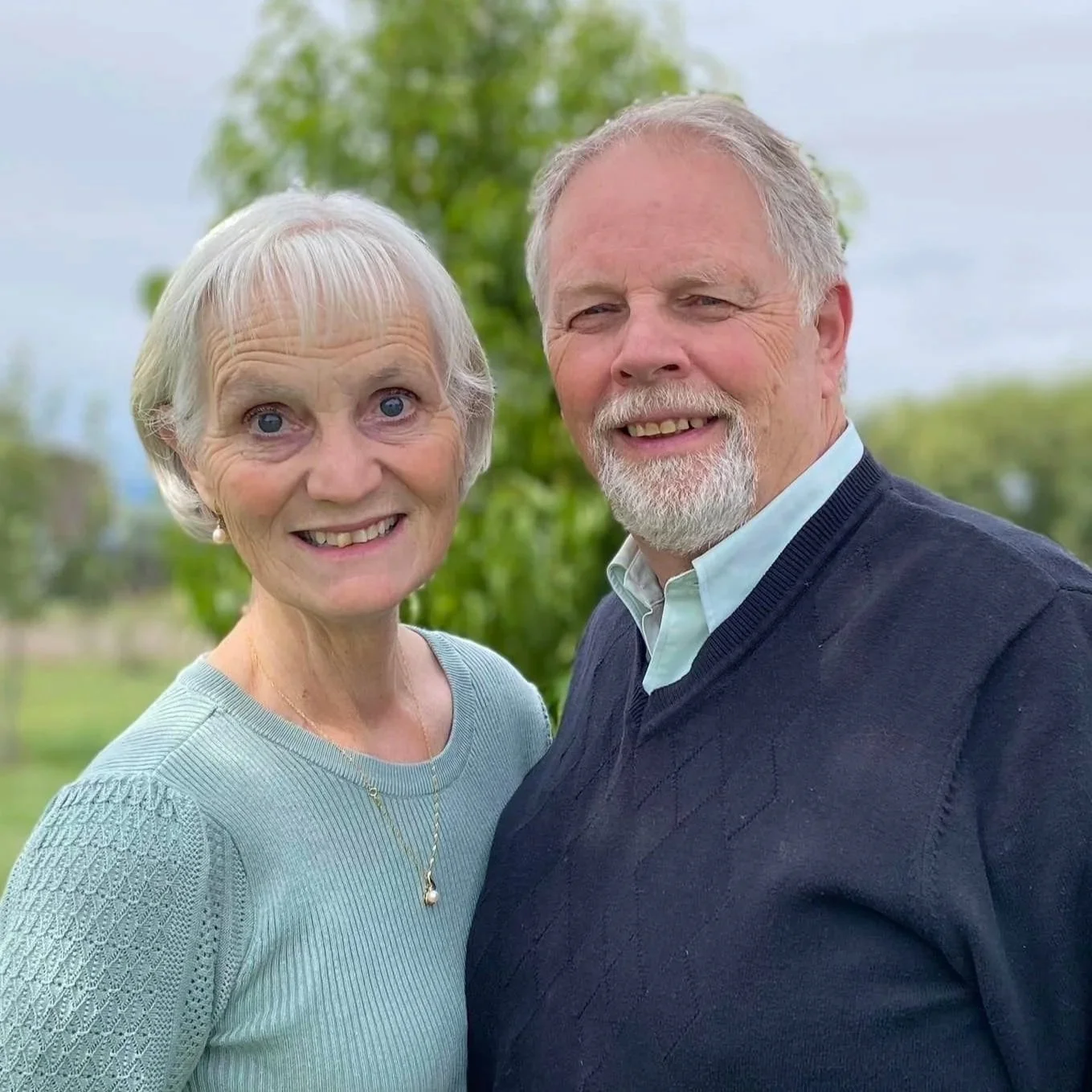 A woman and man standing outside, smiling, with a background of trees and cloudy sky.