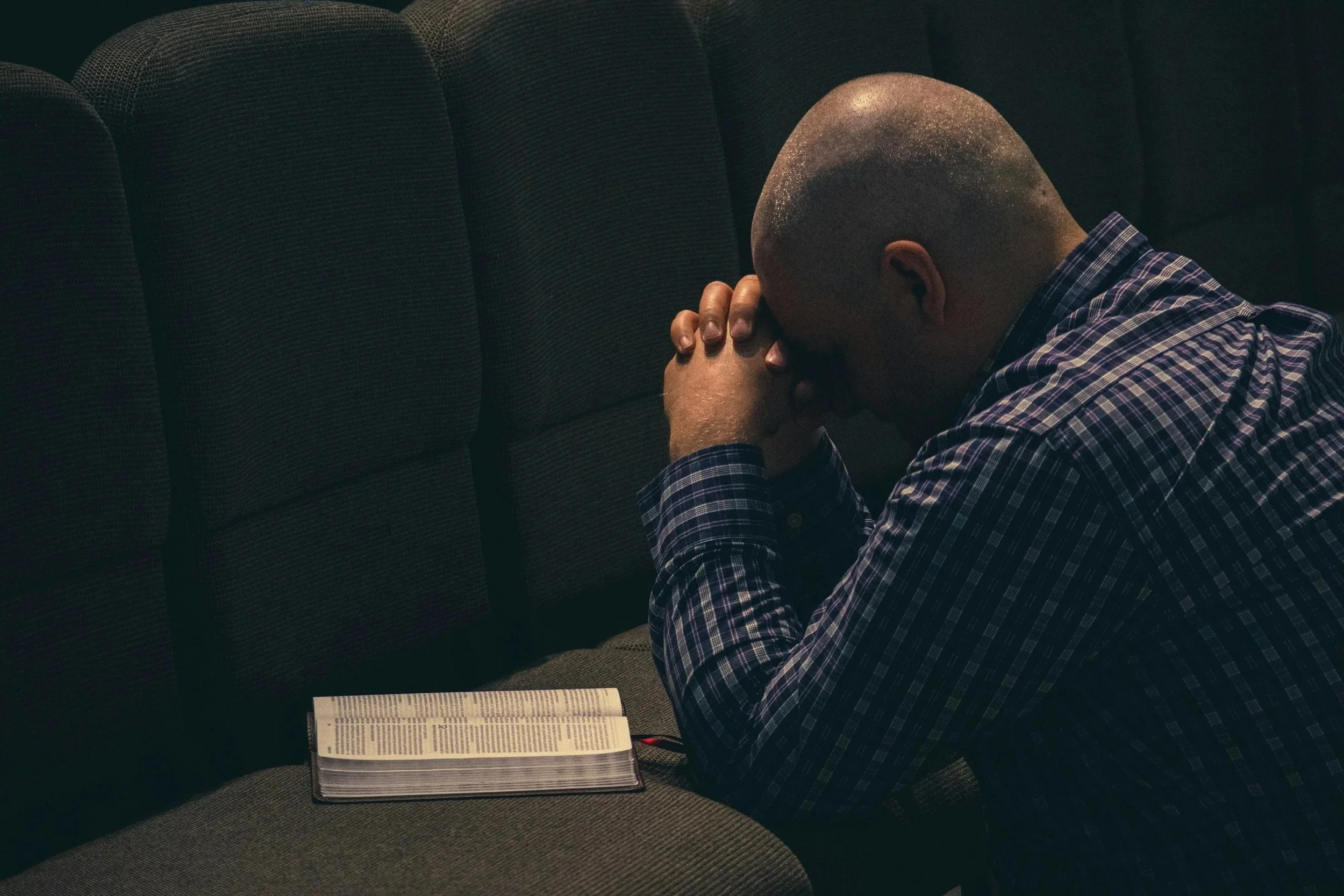 A man with a shaved head sitting on a church pew with his head bowed and hands clasped in prayer, next to an open Bible.