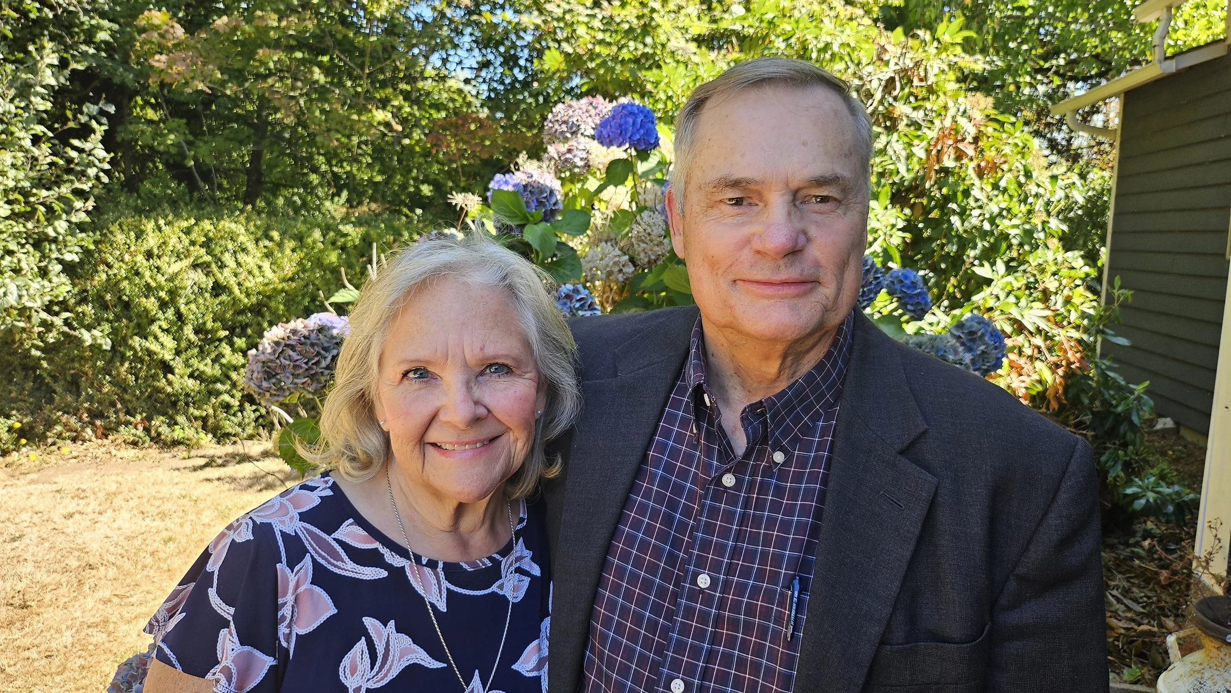 A smiling woman with gray hair and a blue floral dress standing next to a man with gray hair wearing a dark blazer and checkered shirt, outdoors in a garden with green bushes and purple hydrangea flowers in the background.