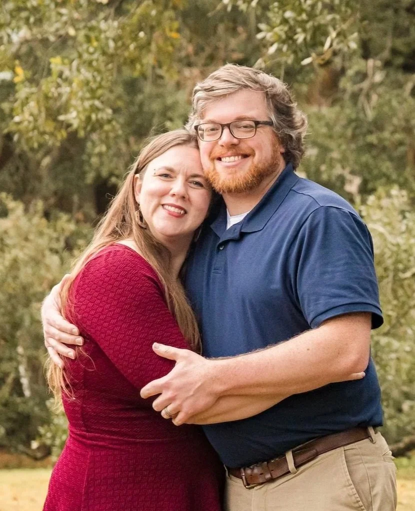 A smiling couple hugging outdoors with trees in the background.