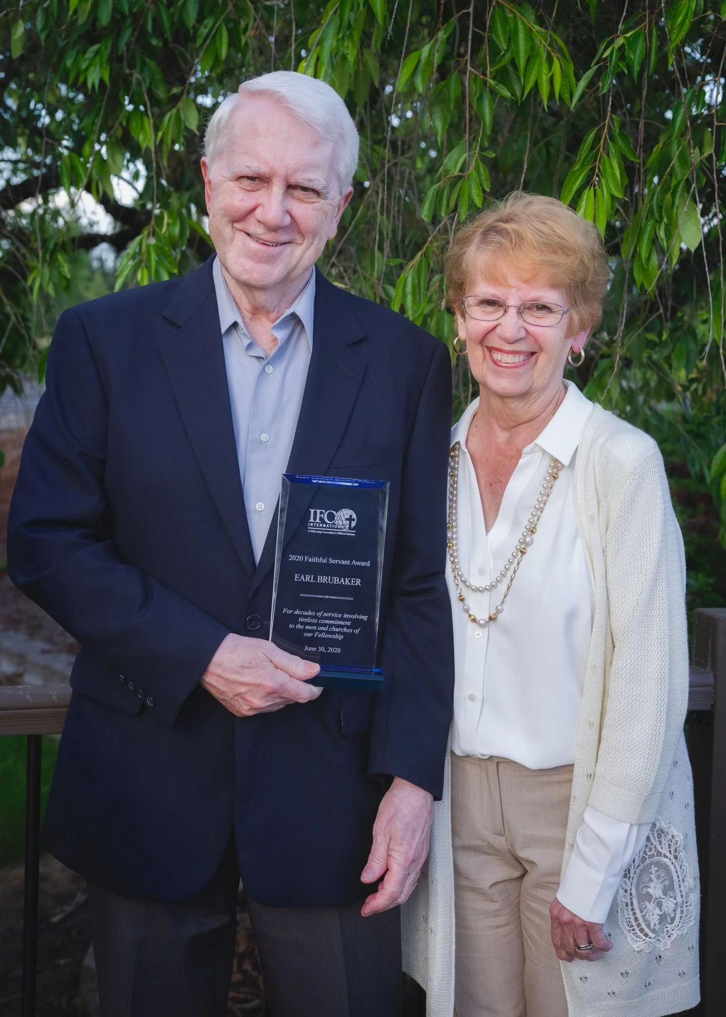 Earl and Shirley Brubaker outdoors, with Earl holding a glass award plaque.