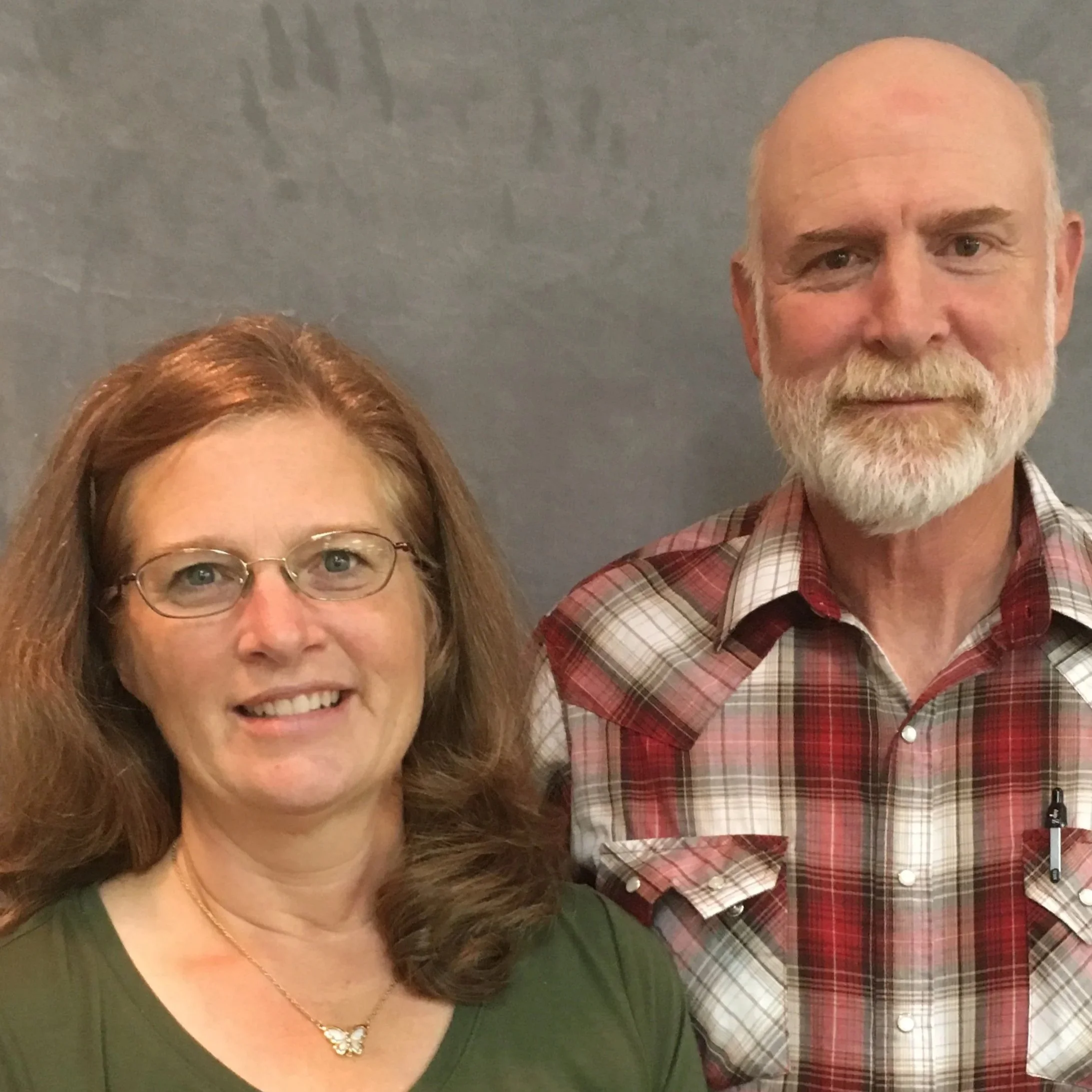 A woman with red hair, glasses, and a green shirt standing next to a man with a white beard, and a checkered red and white shirt, against a plain gray background.