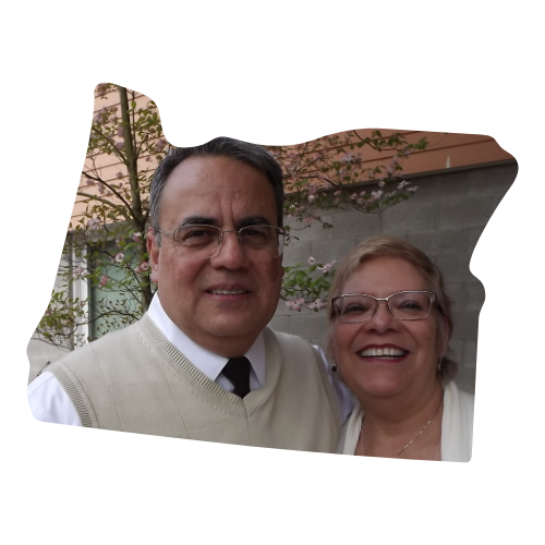 A man and woman smiling outside, with trees and a building in the background.