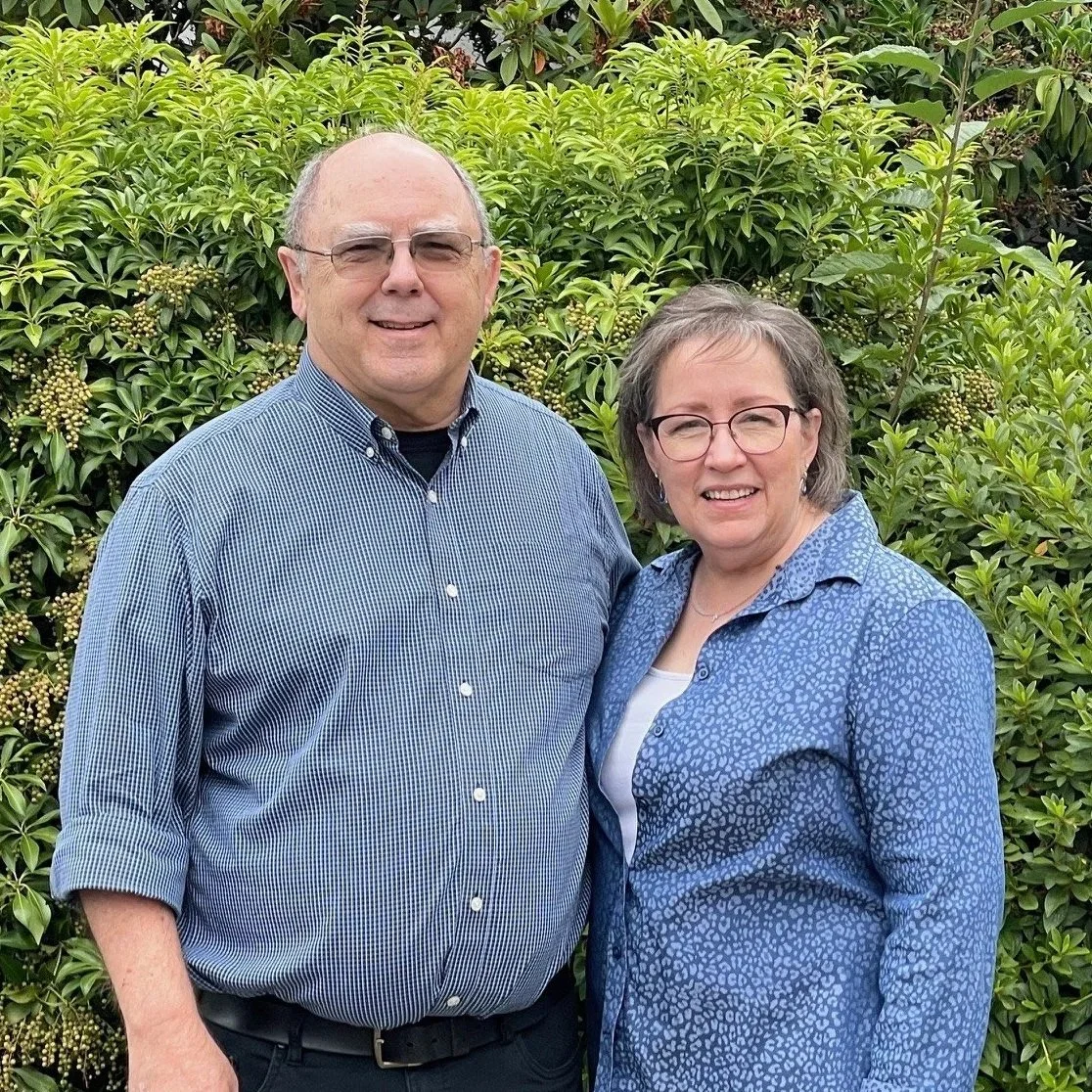 A smiling man and woman standing outdoors in front of green bushes.