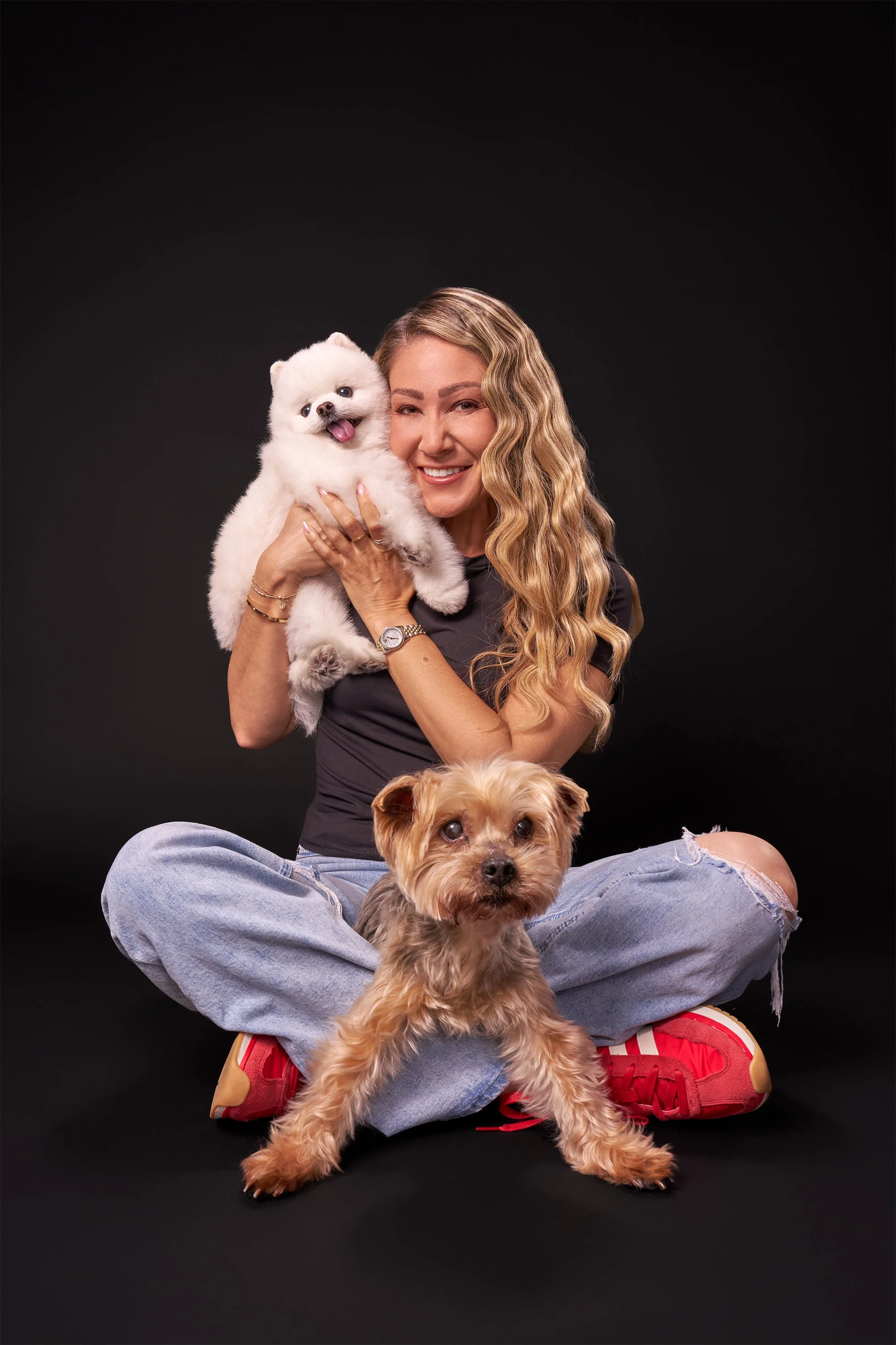 Smiling Latina woman with long hair sitting cross-legged on a black backdrop, holding a yorkie in her lap and a white pomeranian dog in  her arms at pet and family portrait studio in LA