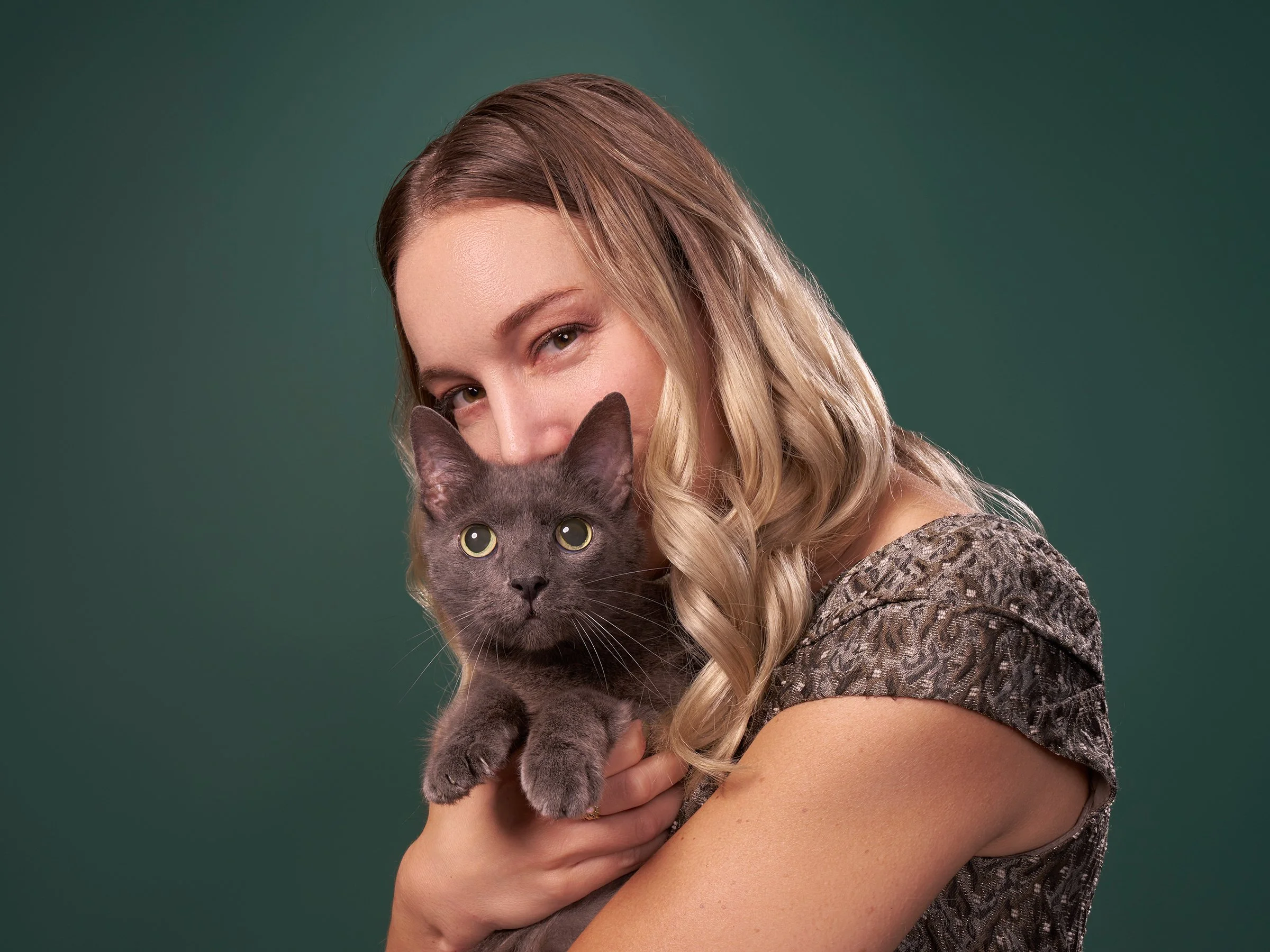 Photo of a blonde woman peering over the head of her gray cat against a green background in Los Angeles pet portrait studio Shuttermutt Photo