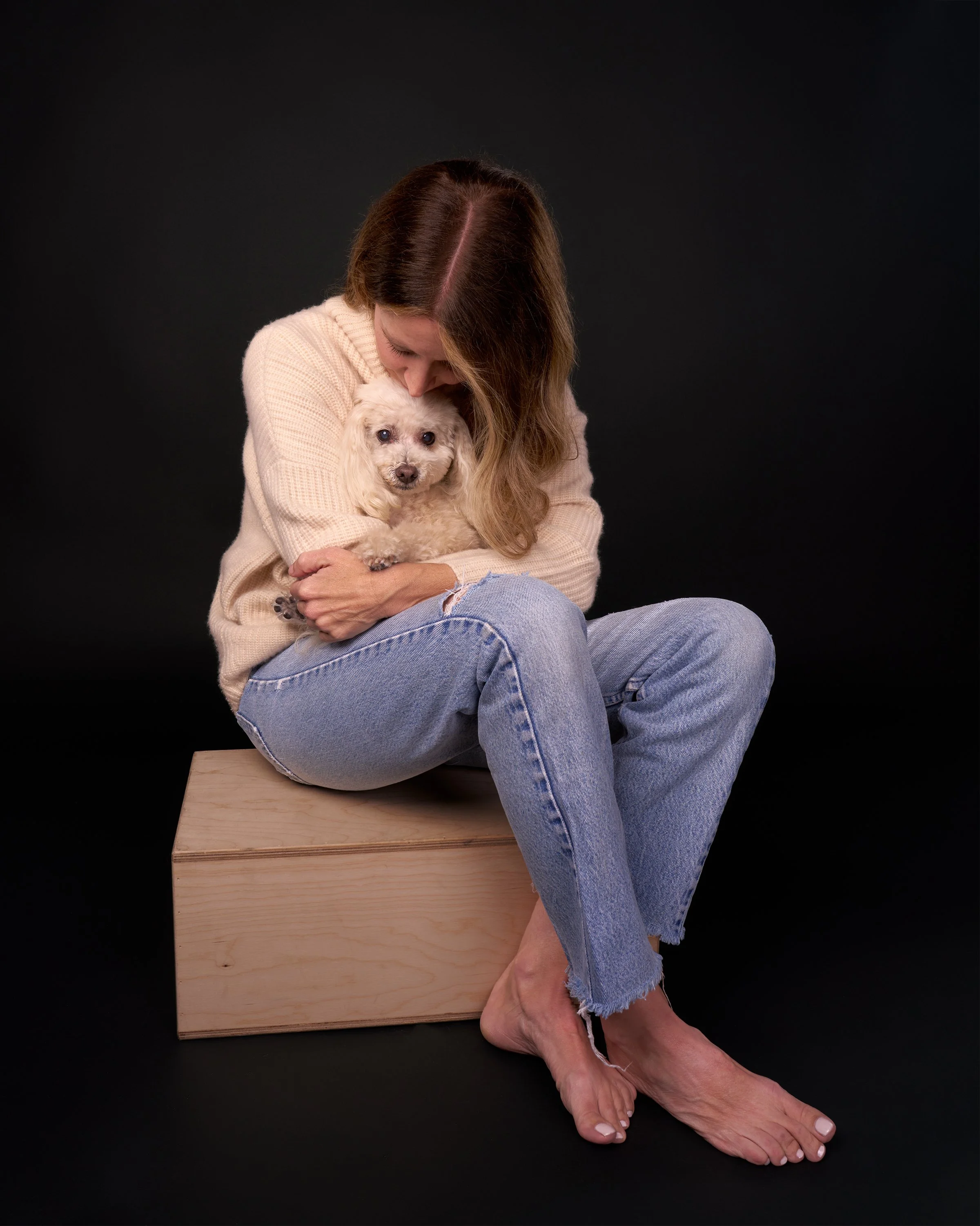 A woman in a white sweater and blue jeans sits on a small wooden box against a black background, leaning her head down to kiss a small white dog sitting in her lap in Los Angeles dog portrait studio