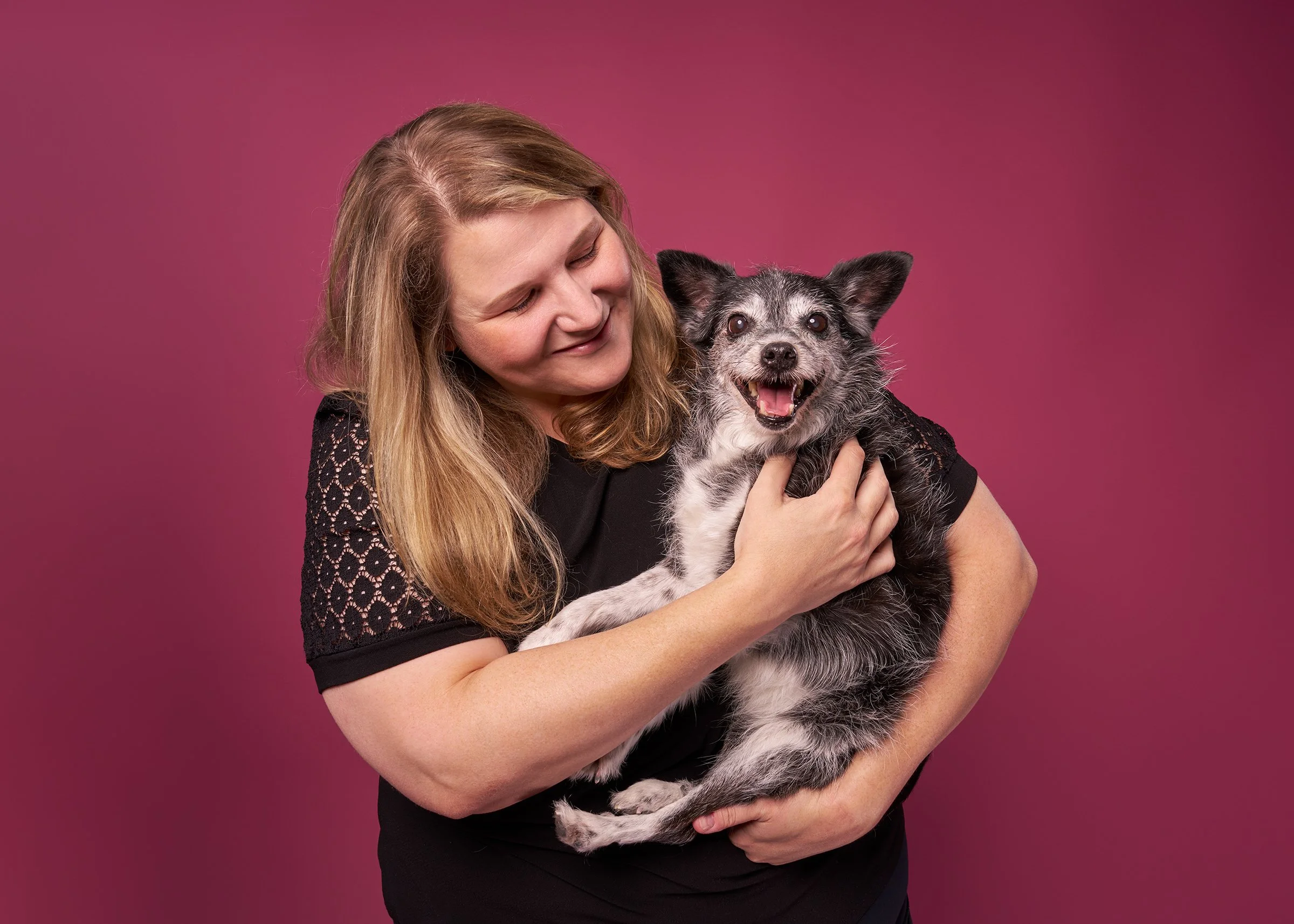 Portrait of a smiling woman holding her smiling black and white terrier dog against a dark pink background in Los Angeles