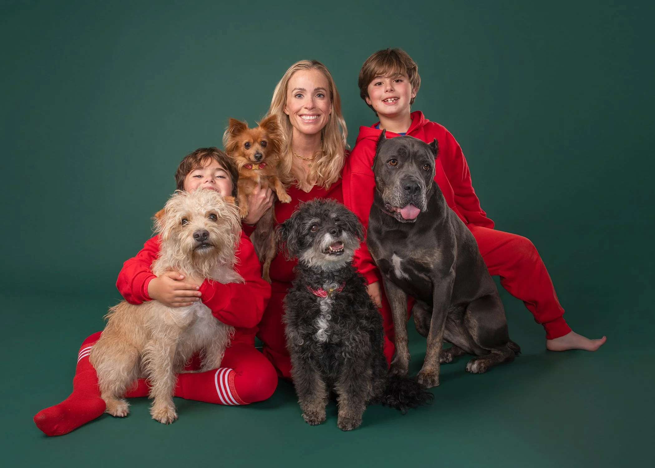 A family in matching red outfits poses on a green backdrop with their four dogs in Los Angeles pet portrait studio Shuttermutt Photo