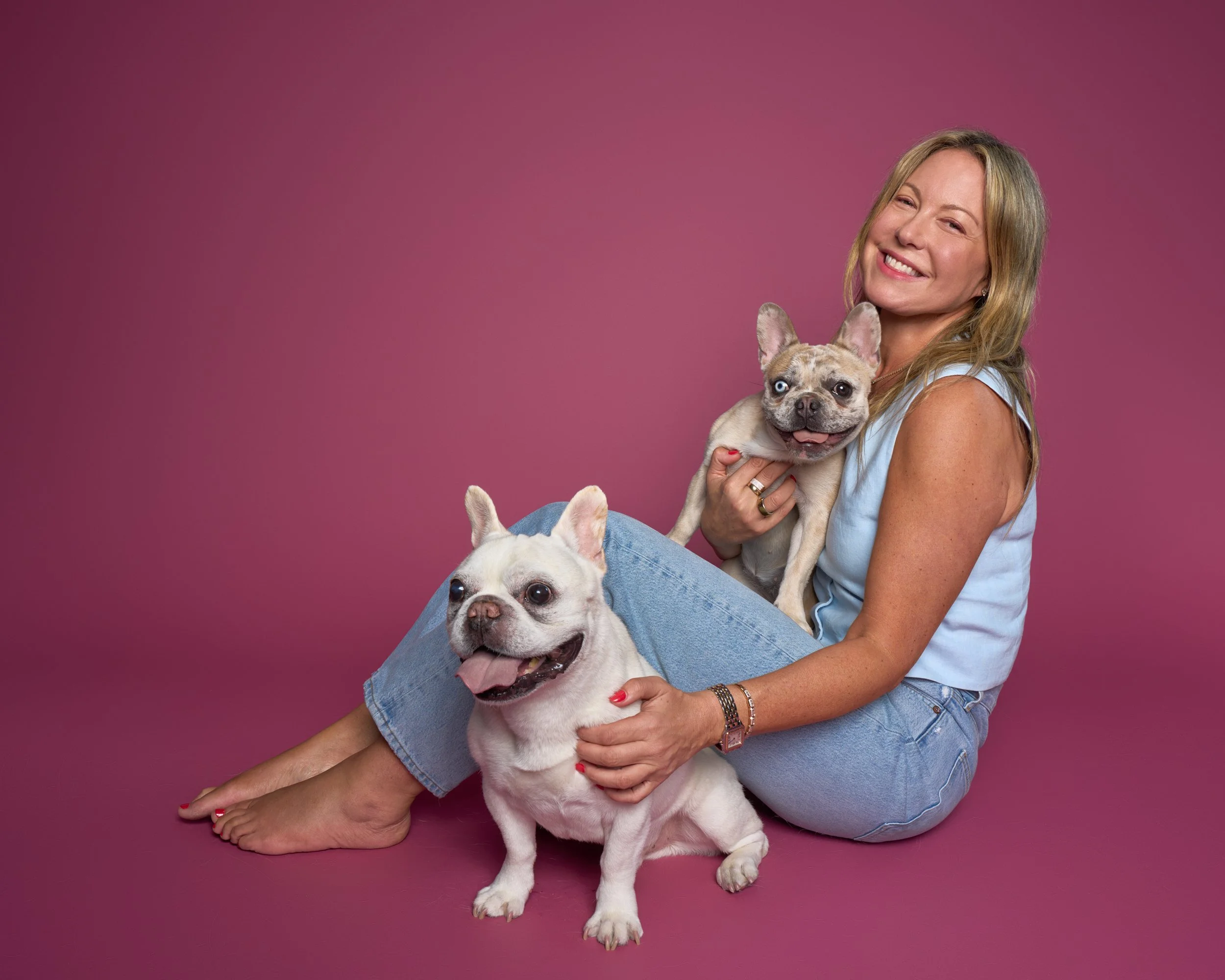 Portrait of a woman smiling and sitting with her two French bulldogs on a pink background in Los Angeles