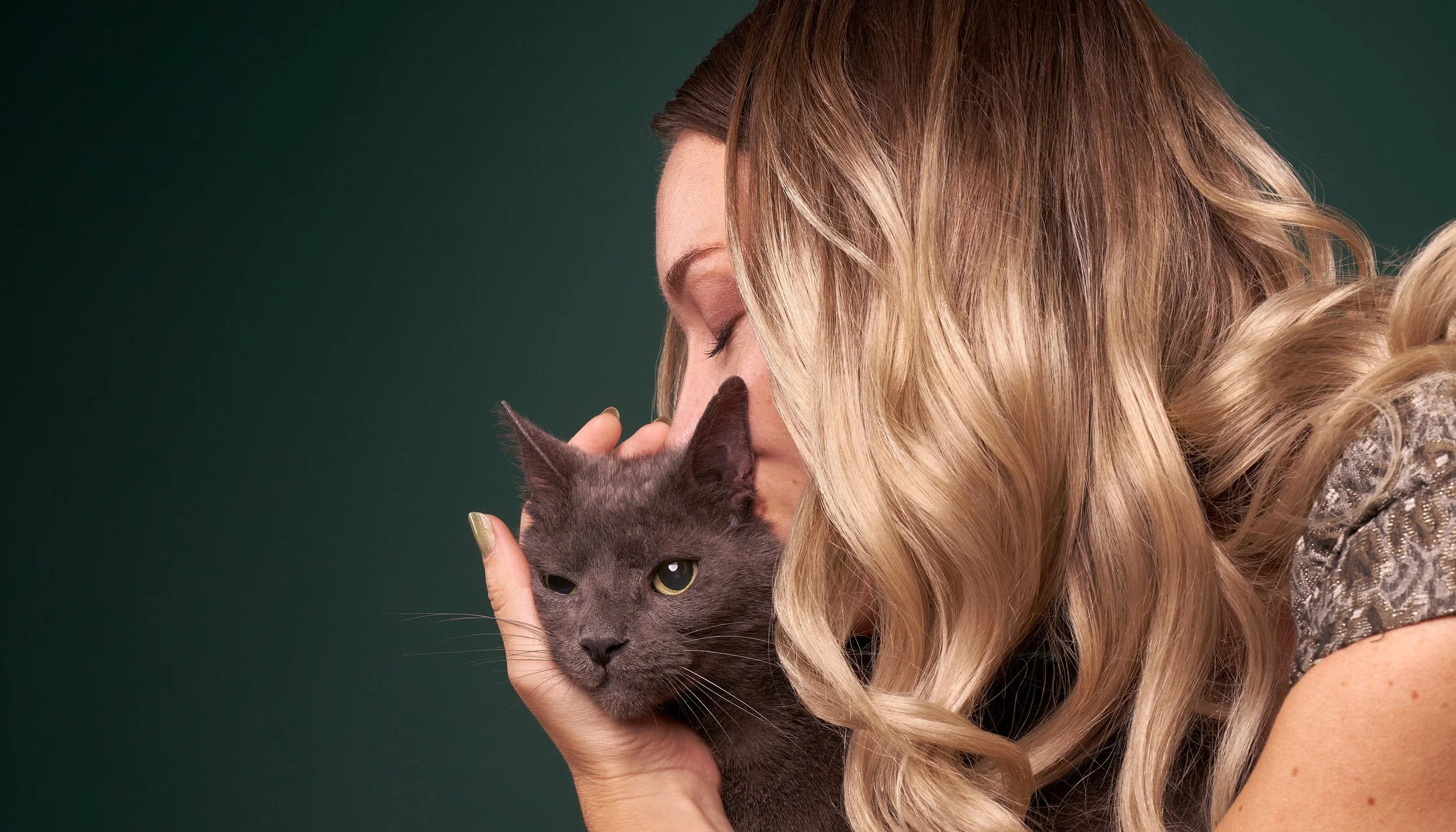 Photo of a blonde woman sweetly kissing her gray cat's head against a dark green background in Los Angeles pet portrait studio Shuttermutt Photo