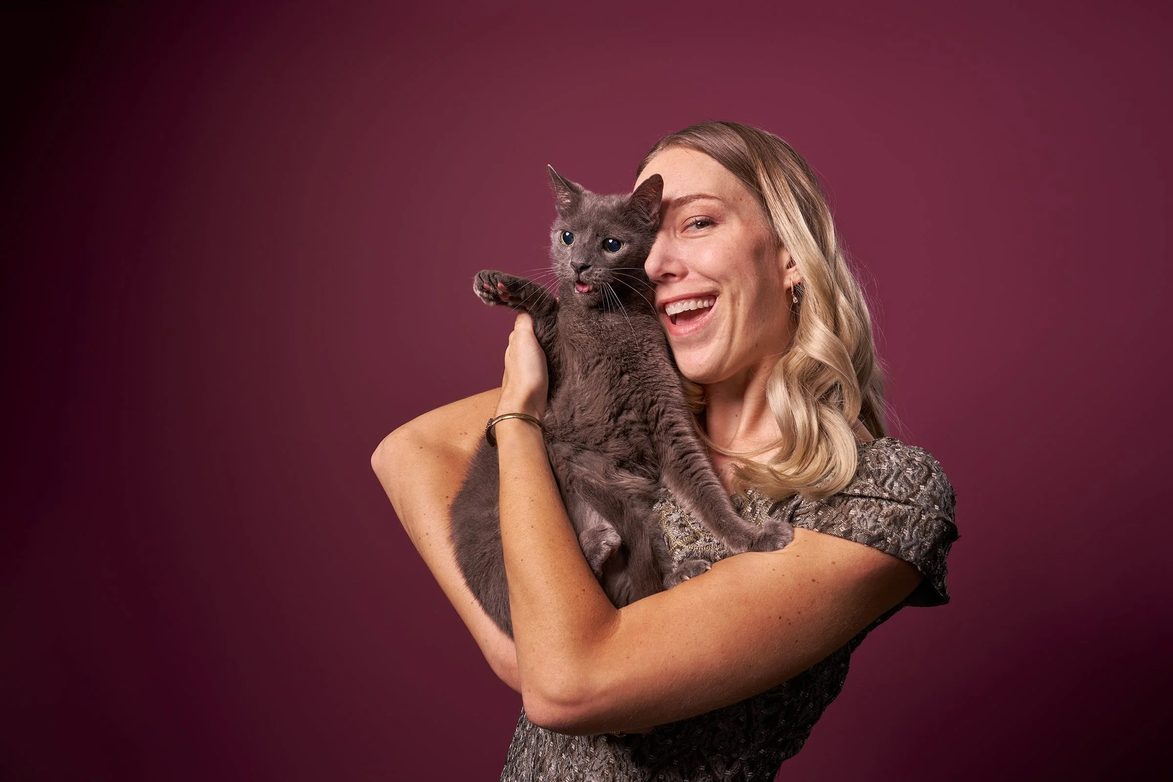 Photo of a blonde woman smiling and holding her gray cat in her arms against a mauve background in Los Angeles pet portrait studio Shuttermutt Photo