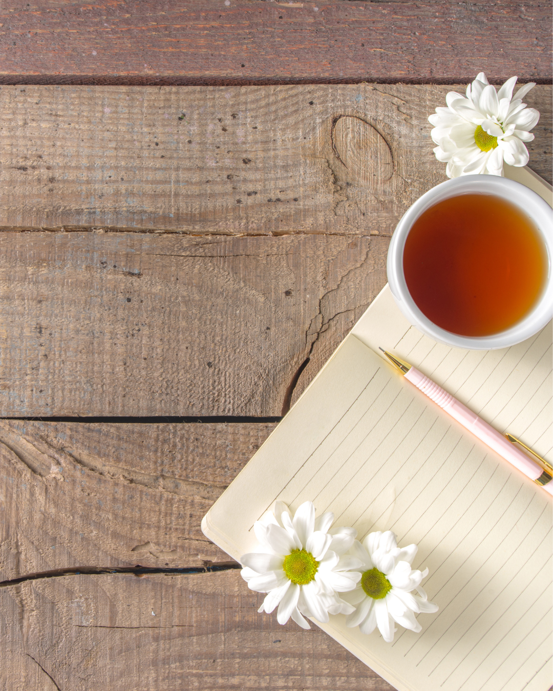 A cup of tea, a notebook, a pink pen, and white daisies on a wooden surface.