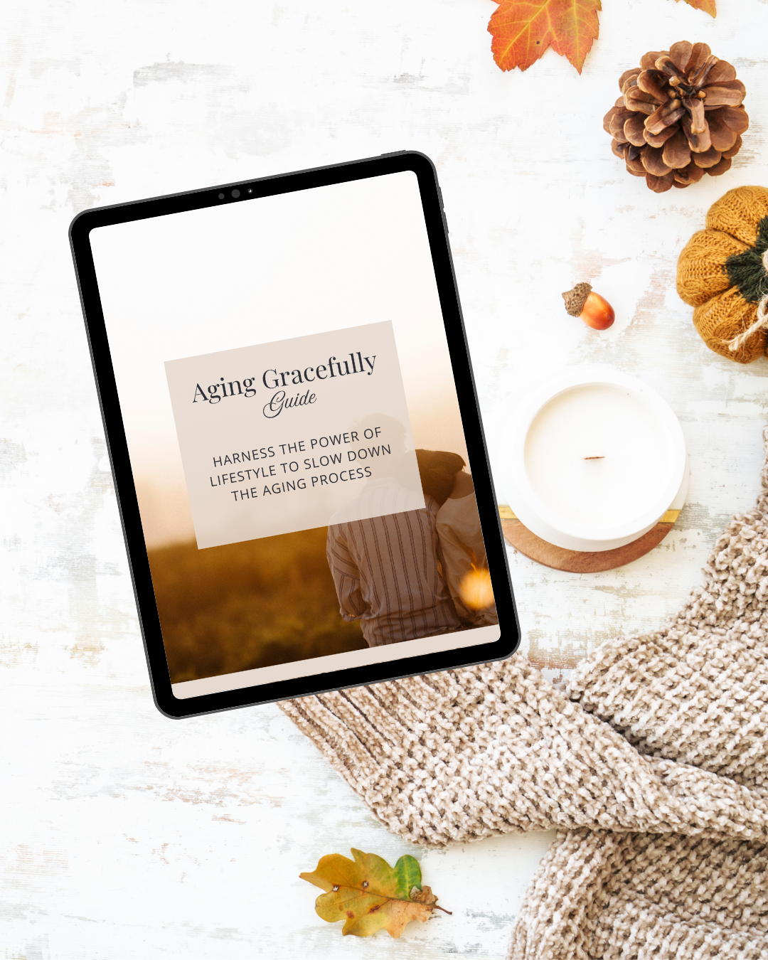 Tablet displaying 'Aging Gracefully Guide' on a white background, surrounded by fall-themed items like pine cones, yellow knitted hat, orange leaves, acorn, candle, and a beige knitted blanket on a white wooden surface.